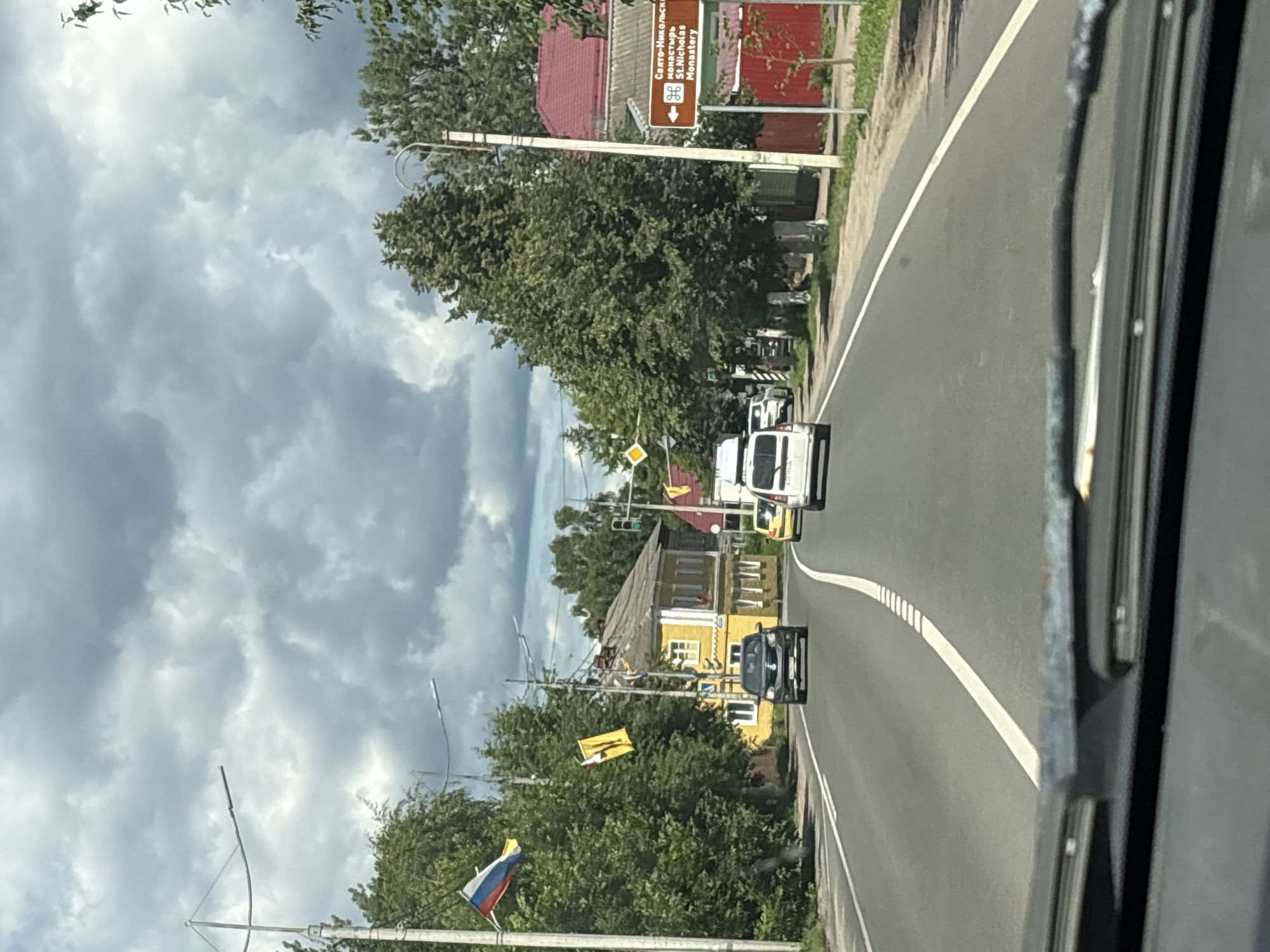 The image shows a view from a vehicle driving on a road in Pereslavl-Zalessky, Russia, under a cloudy sky. On the left side of the road, there are trees and a yellow building. Flags, including the Russian flag, are displayed on poles along the roadside. Several cars are on the road, with one black car in the foreground and a white van and a yellow car further ahead. On the right side of the road, there is a brown sign with text in Russian and English indicating "St. Nicholas Monastery" with an arrow pointing left. There are also trees and a building with a red roof visible on this side. The road has a white dividing line. The perspective suggests the photograph was taken from the driver's or front passenger's seat of a moving vehicle, as evidenced by the dashboard and windshield wipers in the lower part of the frame.