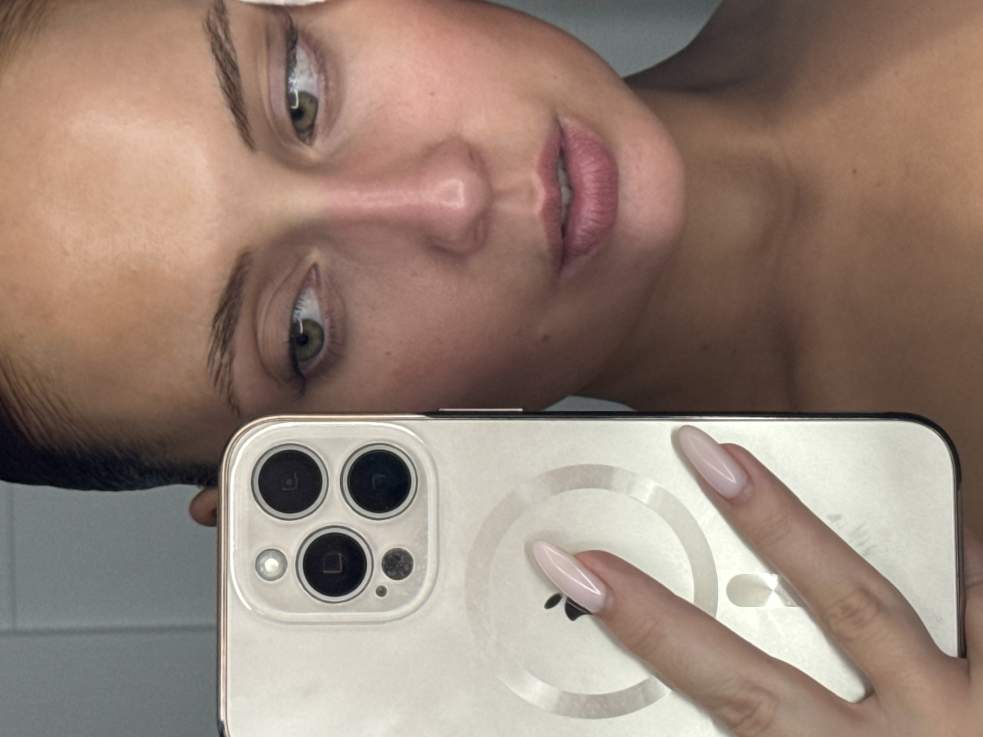 A close-up image captures a person taking a mirror selfie or using a front-facing camera in an indoor setting in Chaikovsky, Russia. The view is angled, showing the upper portion of a person's face from a slightly overhead perspective, including their eyes, nose, and mouth. Their eyes are open with light-colored irises, and their expression is neutral. Dark, defined eyebrows frame their eyes.
A hand, adorned with long, light pink, almond-shaped nails, holds a light-colored smartphone towards the bottom right of the frame. The phone, an iPhone model, features a distinct triple camera setup, a flash, and a circular MagSafe charging ring on its back, visible through a translucent or light-colored case. The Apple logo is discernible beneath the fingers. The person's upper chest and shoulder are also visible in the background, which is a plain, light surface, possibly a wall, reflecting the phone. The activity depicted is a self-portrait, likely taken indoors given the consistent lighting.