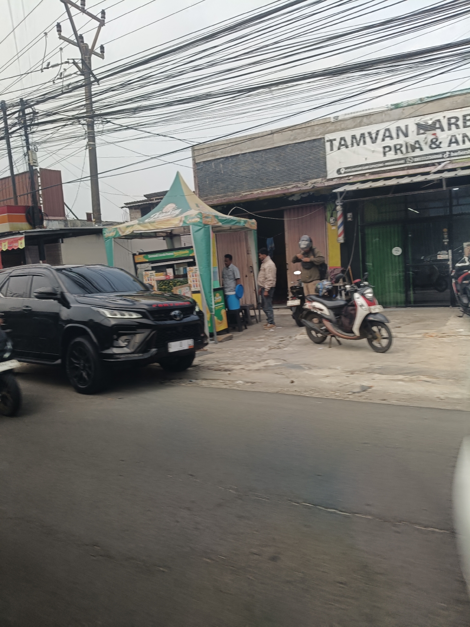 In Saruni, Indonesia, on an overcast day, a black Toyota Fortuner is parked on the right side of a street, angled towards the viewer. The SUV has tinted windows and red "FORTUNER" badging on its hood. To the right of the SUV, a motorcycle with a red and white body is parked, with a person wearing a helmet and jacket standing next to it.
Behind the vehicles, a row of businesses lines the street. One storefront has a banner that reads "TAMVAN BARBER PRIA & AN". Another shop displays a traditional barber pole. A green tent covers a food stall with various items displayed. Several people are visible: two men are standing in front of the barber shop, and another man is near the tent. Electrical wires crisscross above the scene, suspended from utility poles. The street surface is paved and appears somewhat worn.