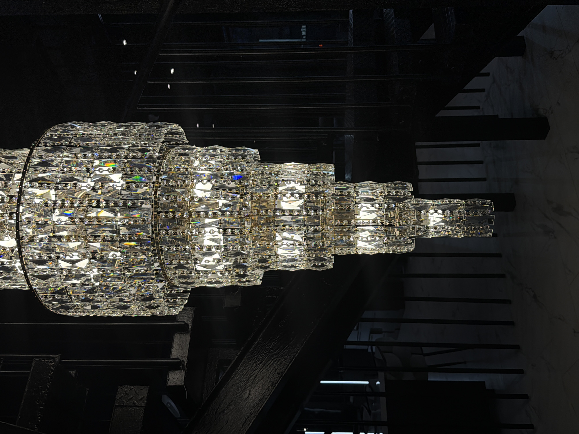 Captured from a low angle indoors, the image showcases a multi-tiered crystal chandelier juxtaposed against a dark metal staircase, with steps that diminish in length on the right. The chandelier, the main subject, is constructed from numerous small, clear crystals, radiating light and reflecting colors. The metal staircase contrasts with the chandelier, with dark, metallic structural elements. The environment is dimly lit, contributing to a dramatic effect. There is no indication of people, actions, or interactions within the image. The location is not discernible from the provided image, therefore the mention of Moscow, Russia is unrelated.