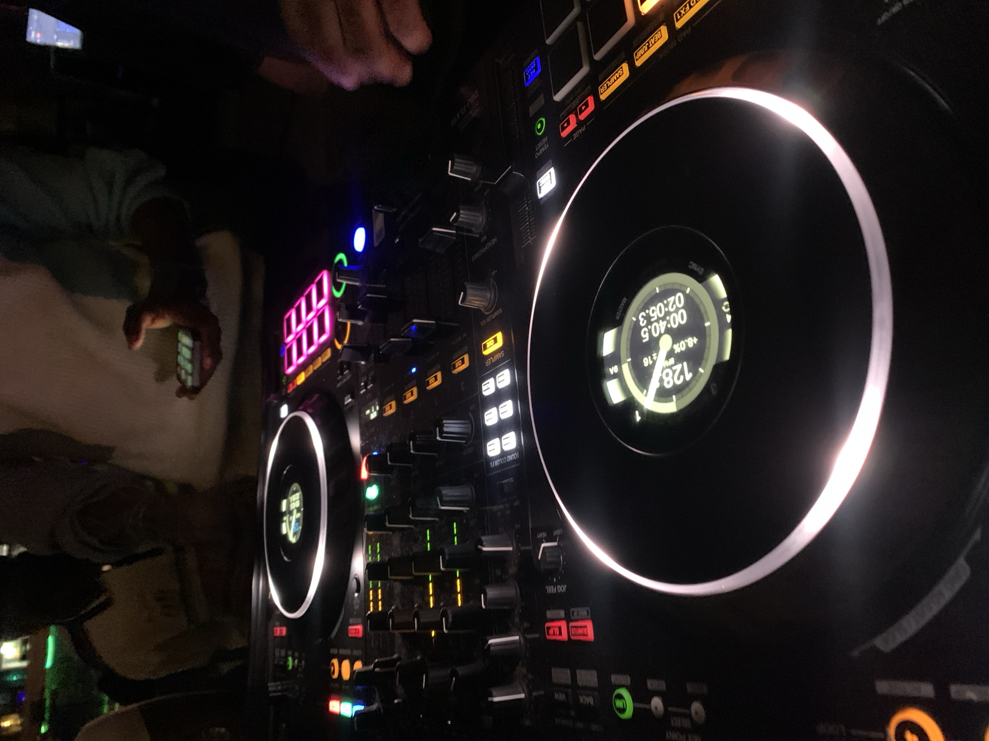 In the dimly lit interior of a club in Howayi, Nigeria, a DJ is seen operating a Pioneer DJM-2000 mixer and two Pioneer CDJ-2000 Nexus turntables. The DJ's hands are visible as they manipulate the controls. The turntables and mixer are illuminated by the colorful lights of the club, with glowing neon pink buttons and a circular display on one of the turntables showing track information. The scene suggests a live DJ set in progress, with the focus on the equipment and the act of mixing music. The background is obscured by darkness, with hints of other patrons and the club's ambiance.