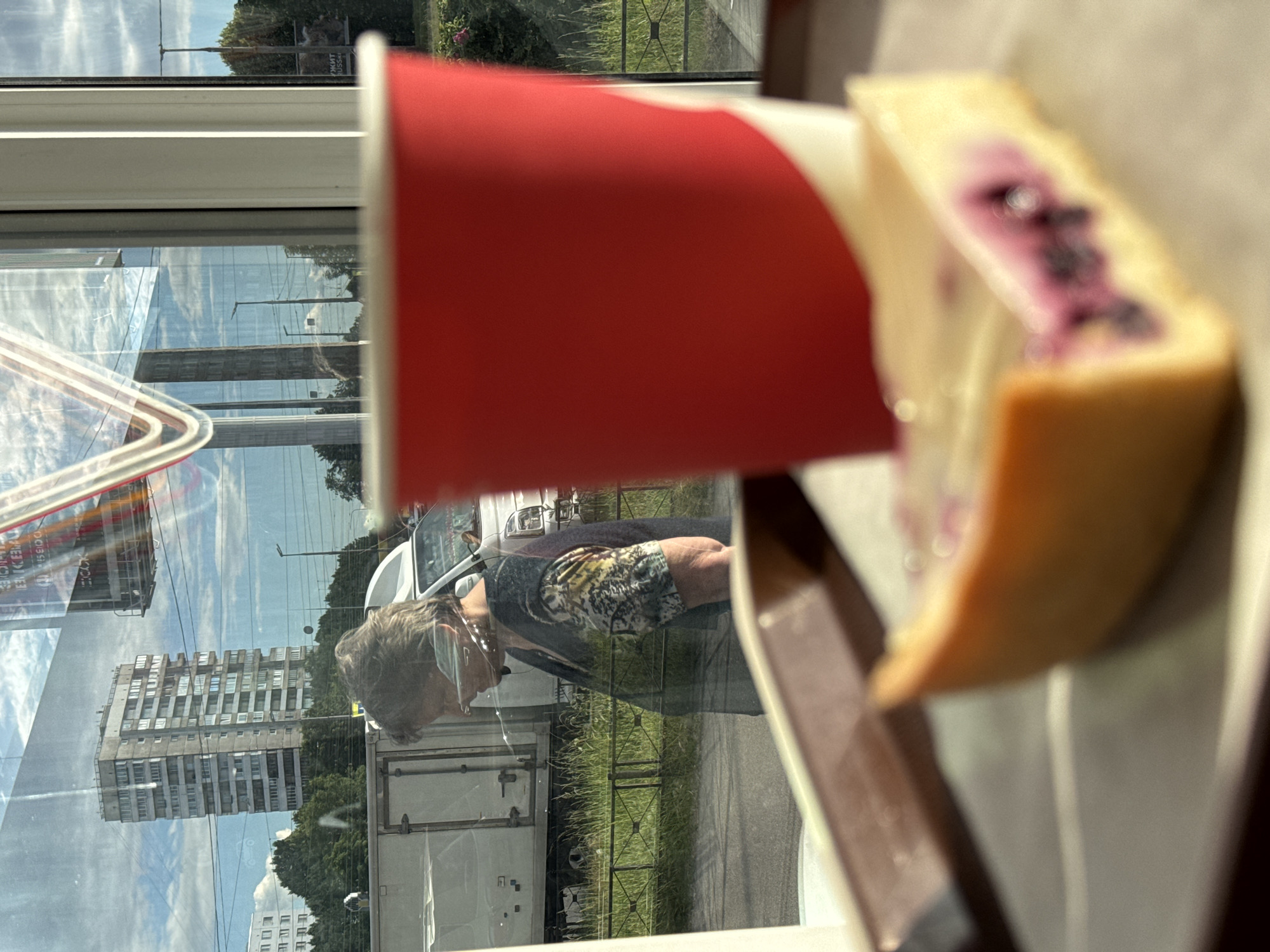 The image shows a slice of cheesecake with blueberries on a white plate, next to a red paper cup. The scene is viewed through a window, and the reflections on the glass are prominent. In the reflection, a person wearing a mask and a shirt with a tiger print is visible. They appear to be looking at something outside.
The background reflection reveals a street scene with apartment buildings, trees, and a cloudy sky, suggesting it is daytime. A white truck and a white car are also visible in the reflection. The presence of the buildings and the overall atmosphere suggests an urban setting, likely in Saint Petersburg, Russia, as indicated by the provided location context. There is no explicit text visible in the image, although some blurry text may be present on signs in the reflection. The overall mood is casual and observational, capturing a moment of everyday life.