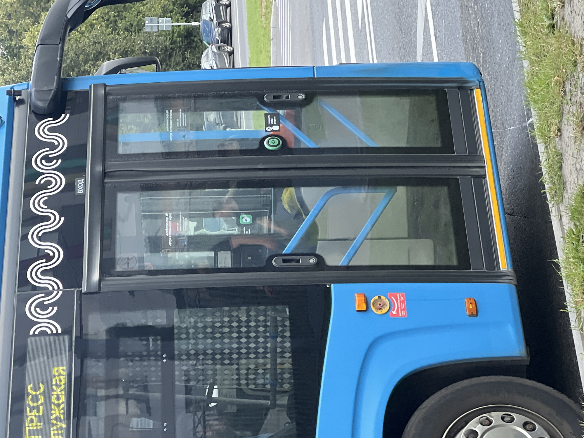 The image shows the side of a blue public bus with its doors open, parked on a street. The bus has a digital display at the top that appears to show "ПРЕСС" and "ПУЖСКАЯ" in yellow text, likely indicating a destination or route. A white wavy design is also present above the doors. A sign inside the bus near the door reads "ВХОД", meaning "Entrance". There are two sets of double glass doors, with one of the doors slightly ajar. A green button with a white figure is visible on the door, indicating a button to open the door. Below this button is a red sticker with white text and an icon. Passengers can be seen through the glass doors, though their faces are not clearly discernible. Outside the bus, there is a crosswalk with white stripes, and further in the background, cars are parked along the roadside. Trees and foliage are visible beyond the cars, suggesting an outdoor urban environment. The overall scene depicts a common urban streetscape in Moscow, Russia, with a bus at a stop.