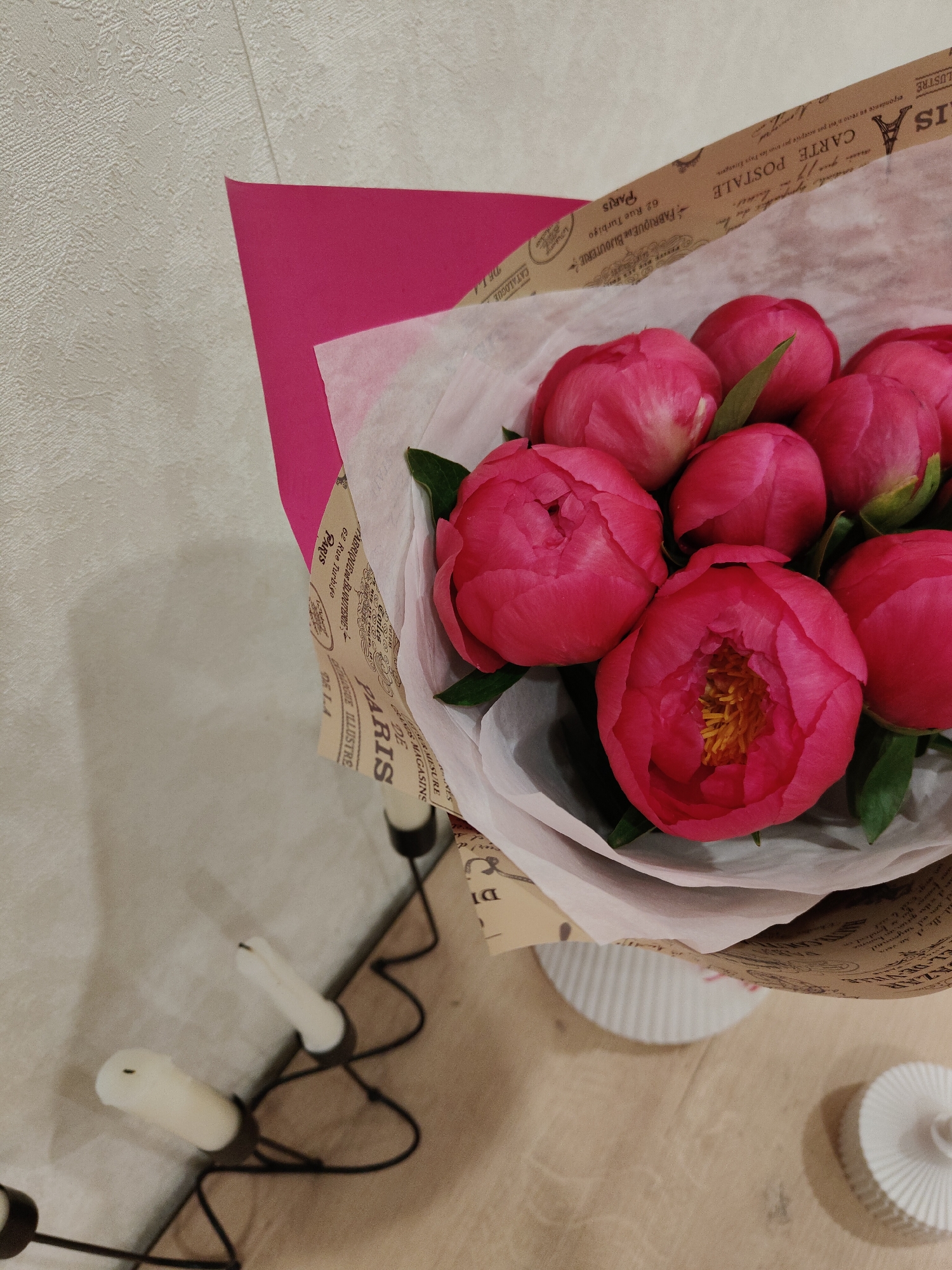 A vibrant bouquet of pink peonies is prominently displayed, set against a light-colored, textured wall and resting on a wooden surface. The bouquet features several full-blooming and partially opened peonies, with one large bloom in the foreground revealing its bright yellow stamens. It is artfully wrapped in three layers: a soft, translucent white paper, a larger sheet of brown paper adorned with French text and decorative motifs, and a solid sheet of bright fuchsia-pink paper beneath.
Visible text on the brown wrapping paper includes phrases such as "CARTE POSTALE" (postcard), "correspondance au recto n'est pas acceptée par tous les Pays Étrangers" (correspondence on the front is not accepted by all foreign countries), "FABRIQUE DE BIJOUTERIE" (jewelry factory), "62 Rue Turbigo," and "PARIS." Other words like "CATALOGUE," "LUSTRE," and "ILLUSTRE" are also discernible, along with a partial number "83110" and other French words and names, suggesting a vintage Parisian theme.
To the left of the bouquet, a black, decorative candelabra with three unlit white candles is visible. On the wooden surface to the right of the bouquet, a white, ribbed cylindrical object, possibly a stand or a vase, is partially in view, with another similar object blurred in the background. The overall setting appears to be indoors, likely a home or a shop.