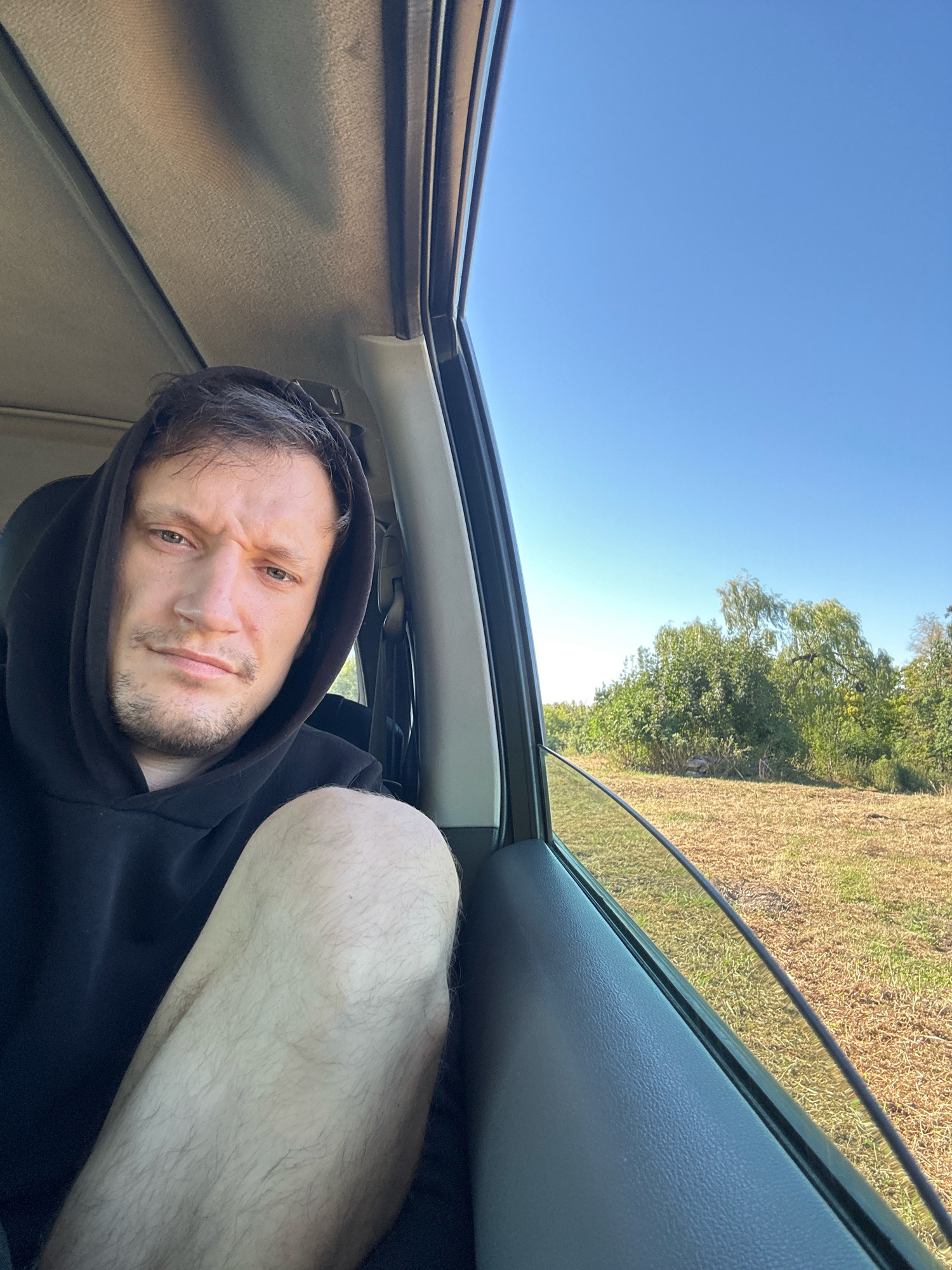 A man with short, dark hair and stubble is captured in a close-up selfie, taken from inside a vehicle. He is wearing a black hoodie with the hood pulled up, partially obscuring his forehead. His left leg is visible in the foreground, resting on the car's interior. The man's gaze is directed towards the camera with a neutral expression. The scene appears to be set outdoors, with a clear blue sky visible through the car window. Beyond the window, a field of dry, light brown grass stretches towards a line of green trees and bushes. The sunlight suggests it is daytime. The specific location of Makiv, Ukraine is not discernible from the visual information provided in the image.