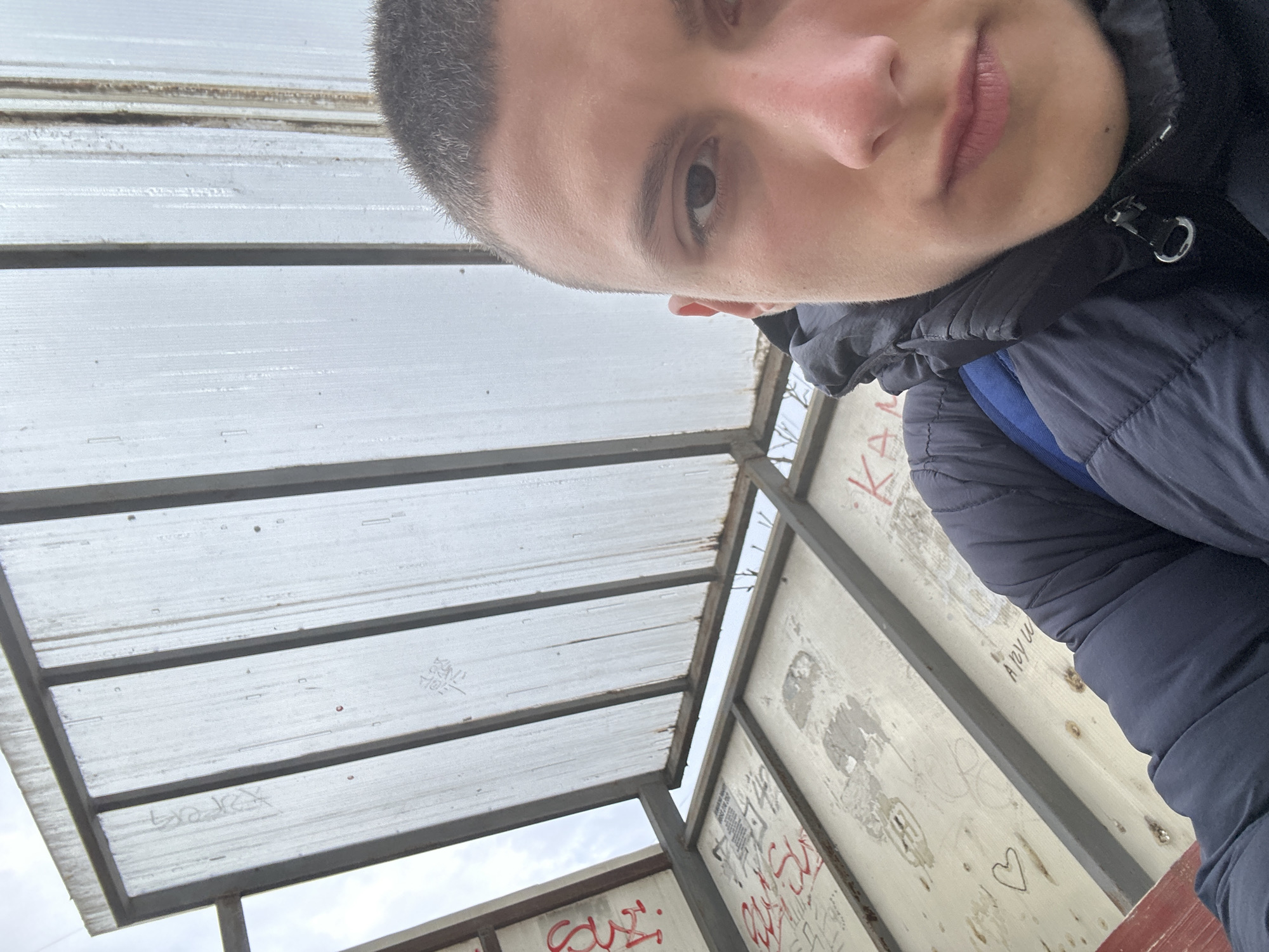 The image is a selfie of a young man with short, dark hair. He is wearing a dark blue puffy jacket. The background appears to be a bus stop or similar shelter with a translucent corrugated plastic roof held up by a metal frame. The walls of the shelter are covered with graffiti. Based on the graffiti and architectural style of the shelter, it's likely the location is Burgas, Bulgaria.