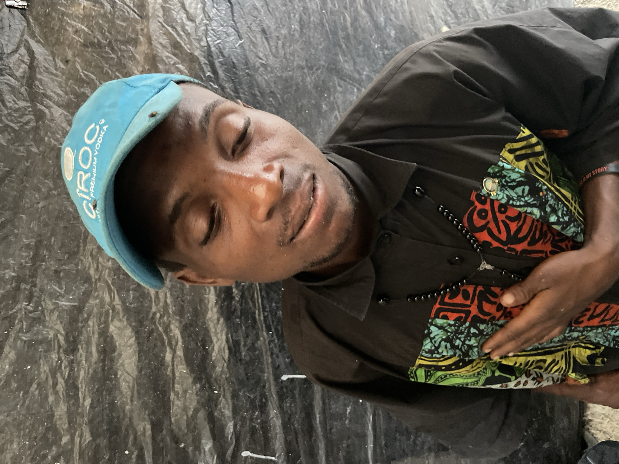 A young Black man wearing a blue cap with "Ciroc Premium Vodka" printed on it and a black shirt with colorful patterns on the front is photographed from the chest up, with his eyes closed and his head tilted down. He has a black beaded necklace with a pendant around his neck, and his left hand is placed over his chest. He is standing in front of a dark, textured background that appears to be a tarp or plastic sheeting. The lighting is even, and the image is in focus. There are no discernible landmarks or environmental features to indicate a specific location within Ilado, Nigeria.