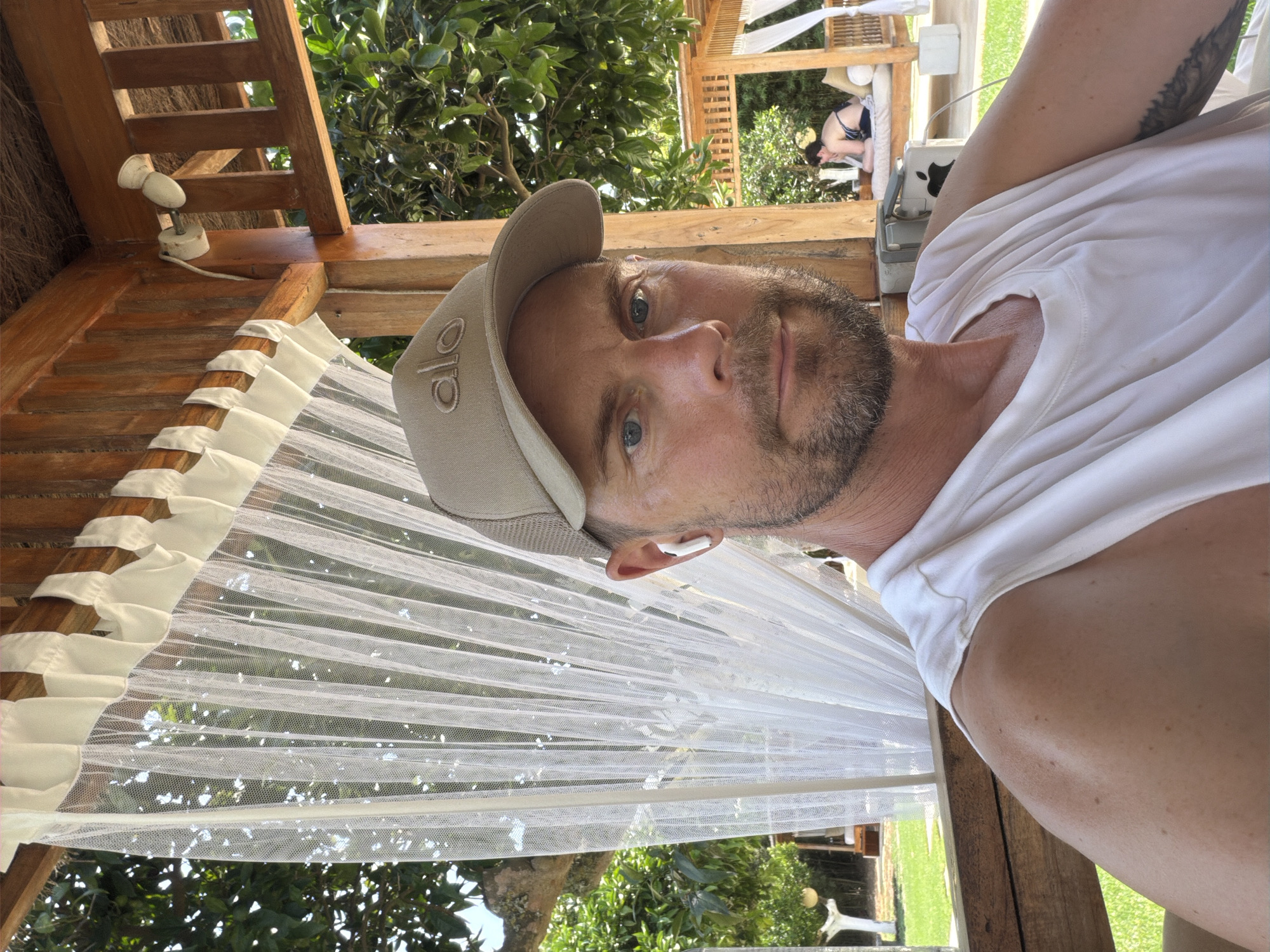 The image shows a man wearing a tan baseball cap and a white sleeveless shirt, posing for a selfie. He has blue eyes and a beard, and he is wearing white earbuds. He is sitting in a wooden structure with a thatched roof, which appears to be an outdoor gazebo or cabana. Behind him, a white sheer curtain hangs, and through it, lush green trees and foliage are visible. In the background, another wooden structure with white cushions and draped fabric can be seen, with a person in a bikini lying on it. The setting suggests a relaxed, possibly resort-like environment, consistent with the city of Sant Joan de Labritja, Spain, known for its natural beauty and tourist accommodations.