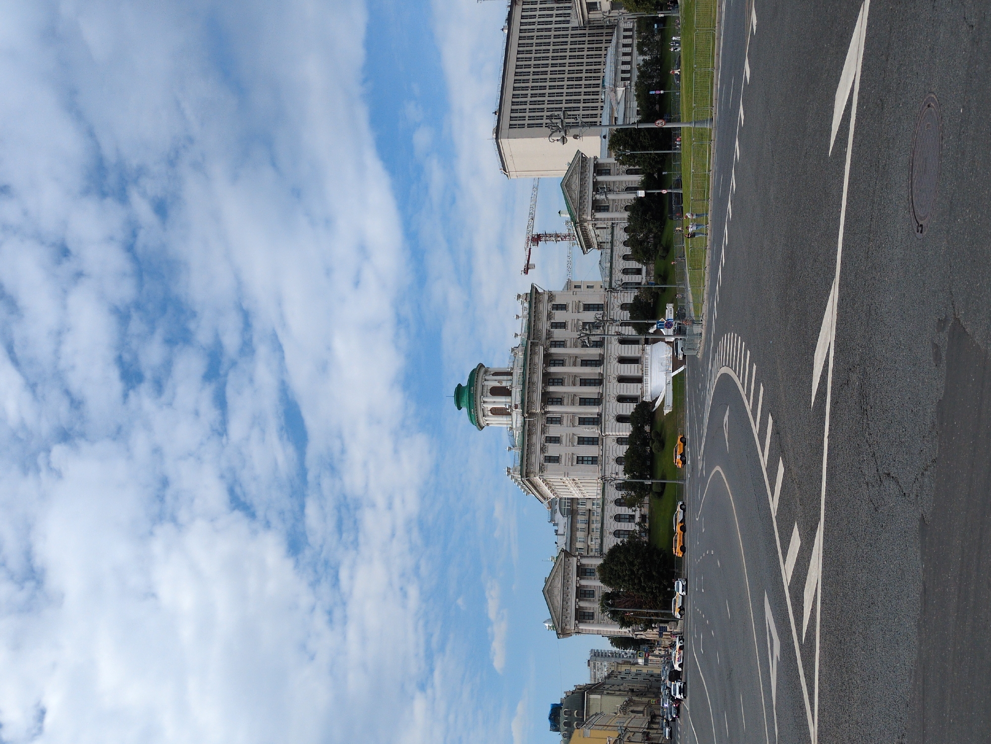 An eye-level, full shot captures a view of an urban intersection with a grand building complex in the background. The foreground features a paved road with clear lane markings, including white arrows indicating direction. Several cars, including what appear to be yellow taxis, are visible on the road.
The background is dominated by a large, ornate building with classical architecture, featuring columns, arched windows, and a distinctive green dome. This building is flanked by other structures, including a modern, rectangular building to the right. A construction crane is also visible behind the buildings. The sky above is a bright blue, dotted with fluffy white clouds. A grassy area with some trees and people can be seen in front of the main building. The overall scene suggests a sunny day in a city.