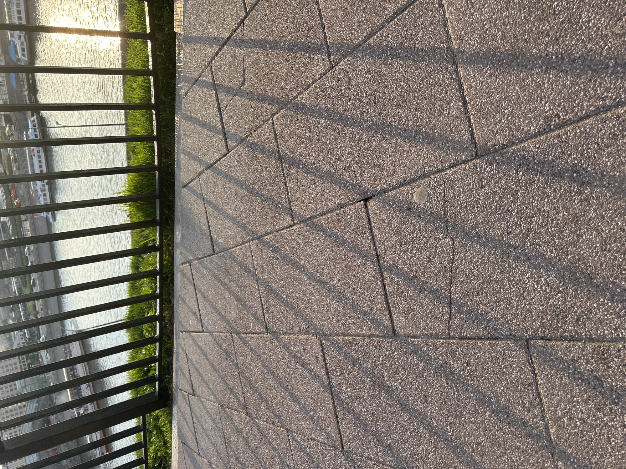 The image shows a close-up view of a paved area next to a metal railing. Beyond the railing, there is grass, water, and buildings in the distance. The paving stones are gray and textured. The shadows of the railing are cast on the paving stones. The sun reflects off the water in the distance. The location appears to be an urban area, possibly a promenade or walkway.