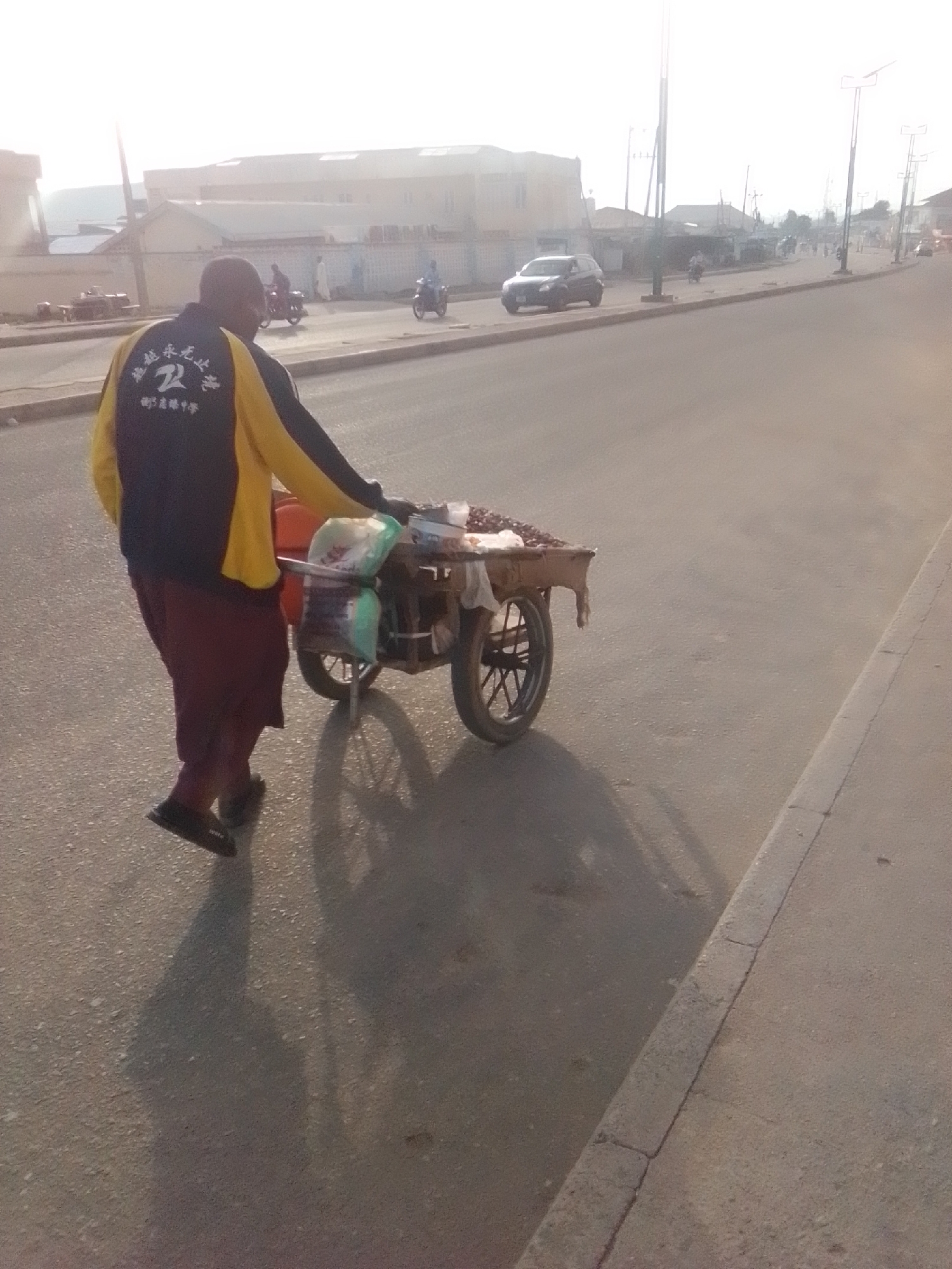 A man walks with a cart on a street in Pantame, Nigeria. The man is wearing a yellow and navy blue jacket with writing on the back and dark red pants. He is pushing a two-wheeled cart that is laden with various items, including bags of what appear to be goods and some sort of produce. The cart has a metal frame and a wooden top. The street is paved, and a sidewalk is visible on the right side of the image. In the background, there are buildings, streetlights, and other vehicles, including cars and motorcycles, suggesting an urban environment. The sky is bright and hazy, indicating either early morning or late afternoon lighting.