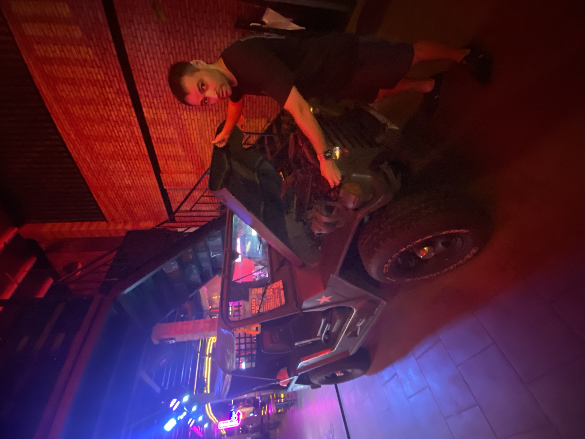 In a dimly lit indoor space with brick walls illuminated by red lighting and neon blue lights in the background, a man in a black t-shirt and shorts stands next to an open-hooded military-style jeep. The jeep is dark colored with a red star on its side. The man is looking towards the camera and has his hands near the engine. The floor is tiled and reflects the colored lights. The overall impression is of a bar or entertainment venue, possibly with a themed decor.
