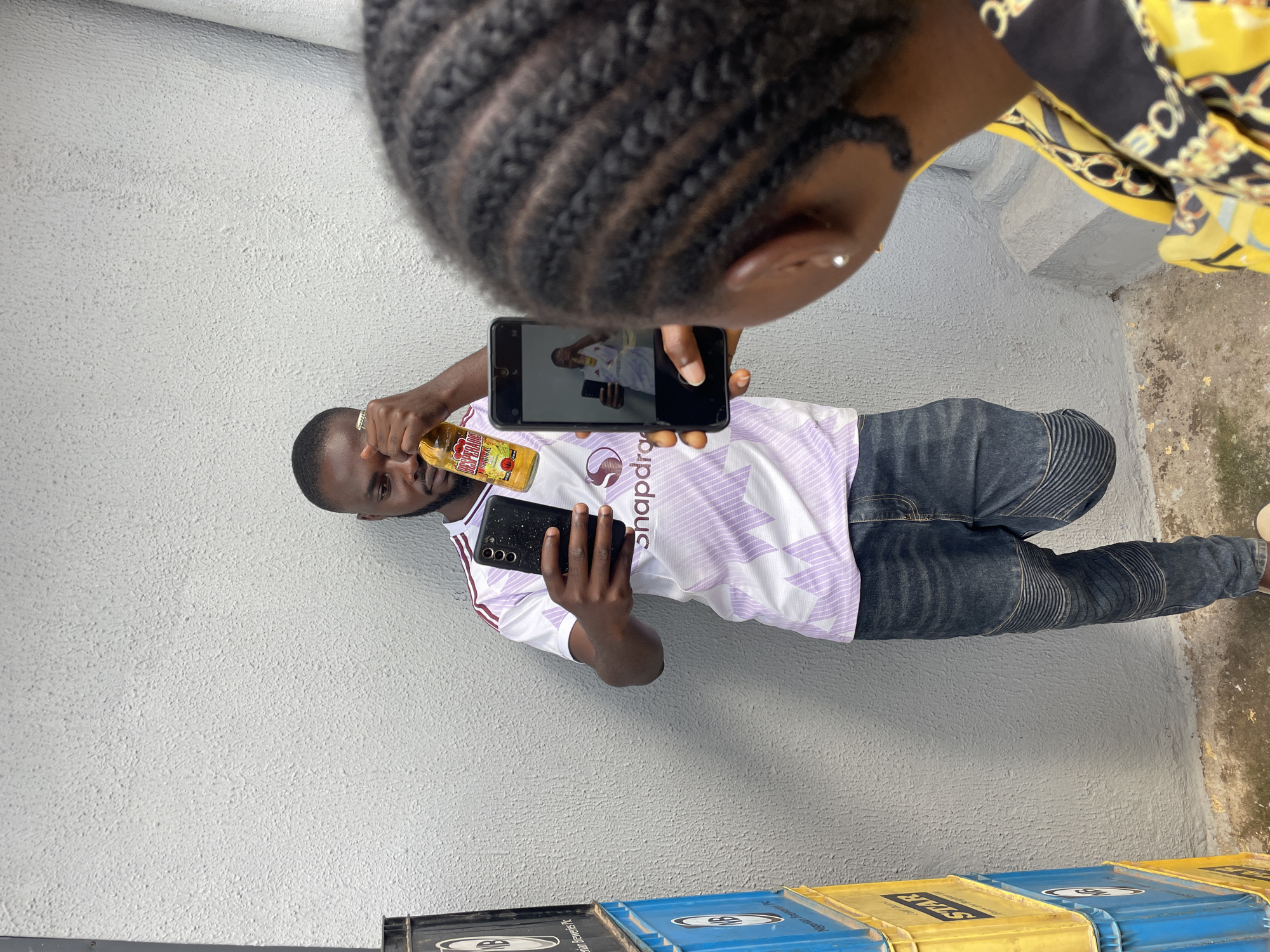The image shows two people and a bottle of beverage. One person, a man, is holding a bottle of "Tango" orange soda and a black smartphone. The man is wearing a white and purple striped t-shirt and dark jeans. He is looking at the smartphone, which is being held by another person from a top-down perspective. The second person, whose face is not fully visible, is holding the smartphone and appears to be taking a picture of the man. The background is a textured grey wall, and the ground appears to be a mix of concrete and dirt. In the lower part of the image, there are some blue and yellow plastic crates. The scene appears to be outdoors, possibly in a public space, given the crates. The lighting suggests it is daytime. The overall mood seems casual and relaxed.