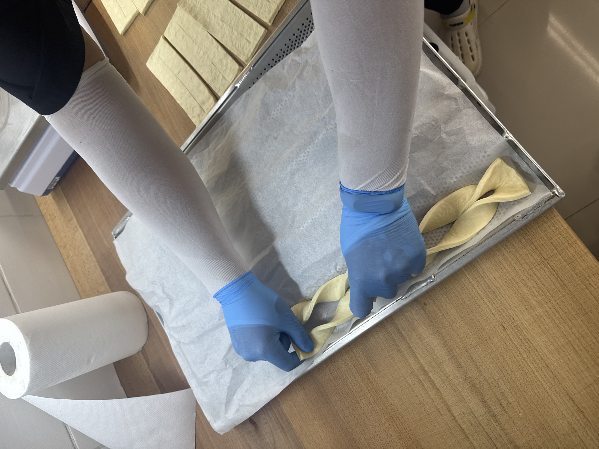 A person wearing blue gloves and white arm sleeves is preparing dough twists on a baking sheet lined with parchment paper. The baking sheet rests on a wooden surface next to a roll of paper towels. A tray with rectangular dough strips sits nearby. A scale can be seen in the background. The location is Vracar Urban Municipality, Serbia.