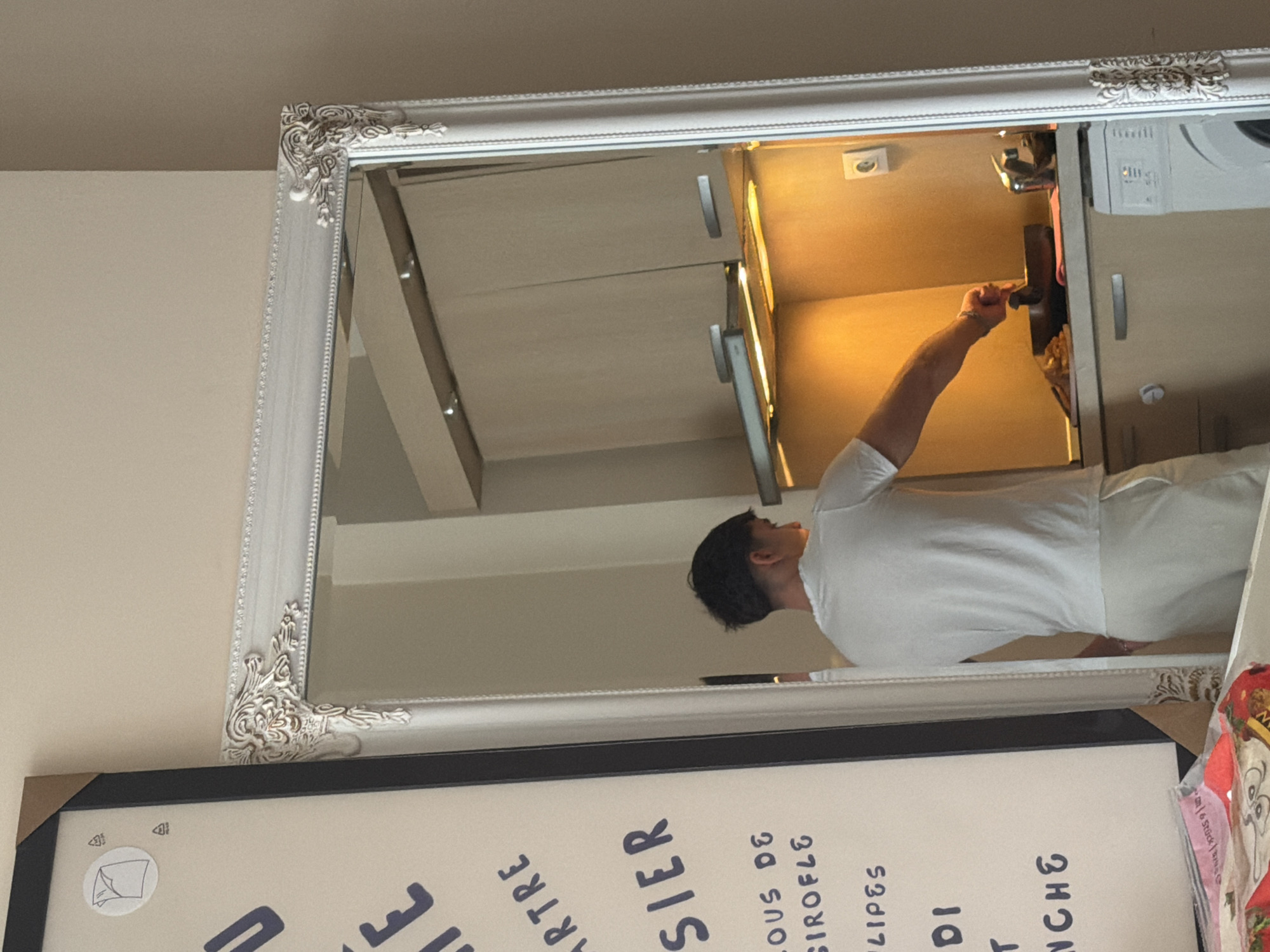 A mirror with a white ornate frame reflects a person opening a cabinet in a kitchen. The kitchen features light-colored cabinets and appliances. Below the mirror is a framed print with French text. The location is likely Sofia, Bulgaria, based on the reflection in the mirror. The person is wearing a white shirt. A packet of goods is located on the right corner of the painting.