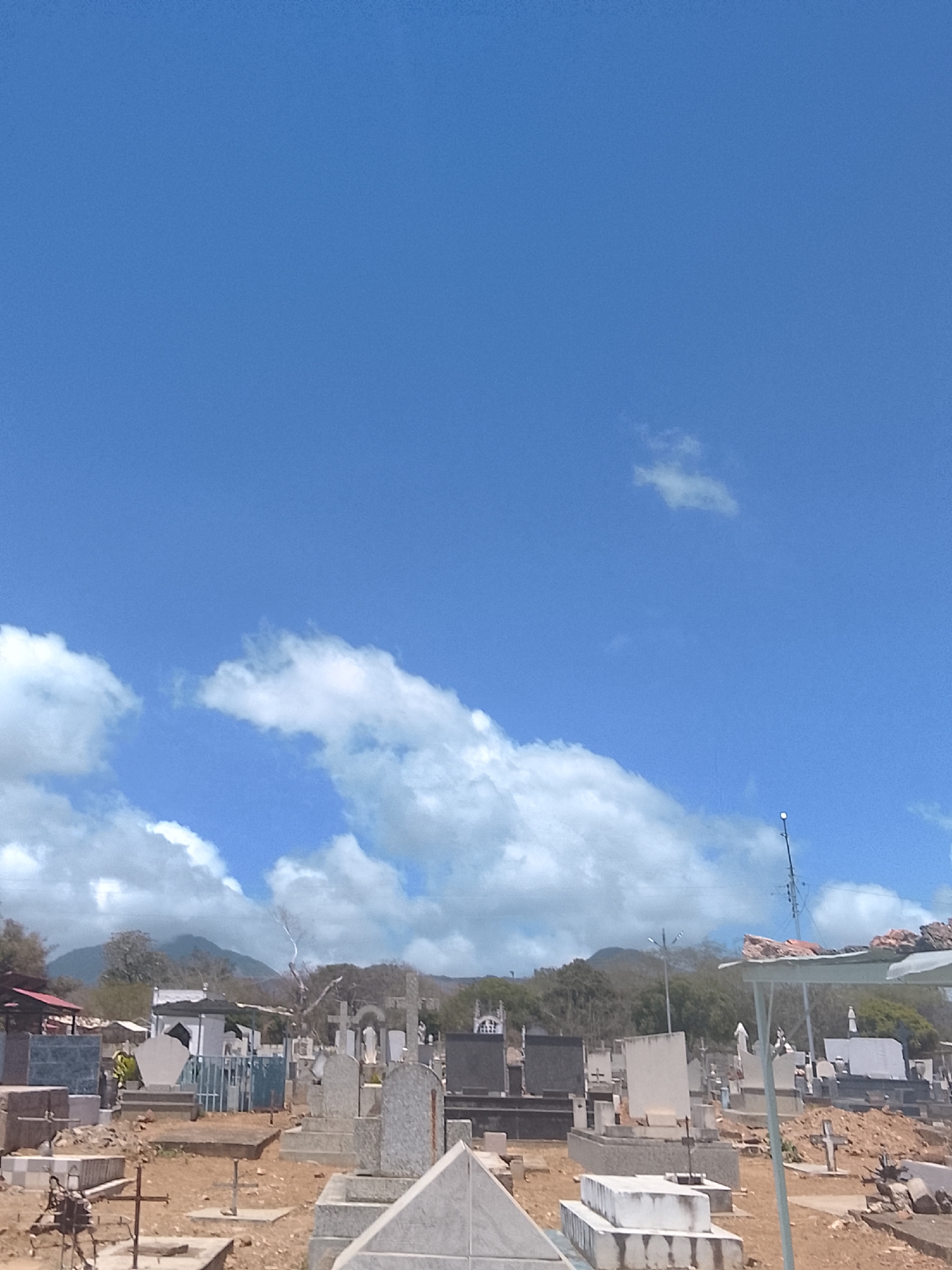 The image depicts a cemetery located in Altagracia, Venezuela. The foreground features numerous tombstones and graves, varying in style and condition. The ground is dry and appears to be a mix of dirt and gravel. In the background, there is a treeline and a mountain range under a partly cloudy blue sky. The overall scene appears to be taken during daylight hours with clear visibility. A utility pole extends from the right side. The general impression is of a well-used, old cemetery.