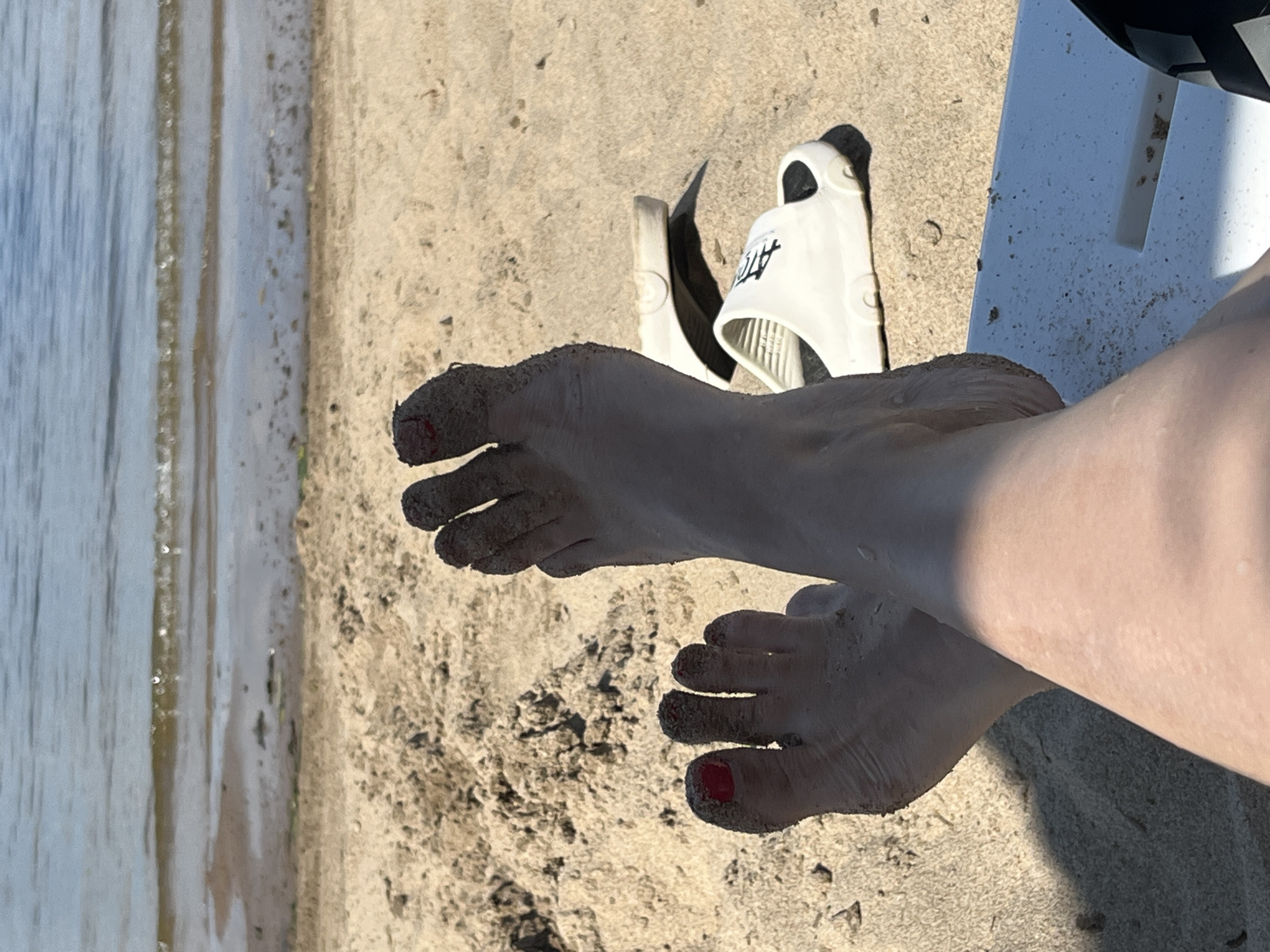 A first-person perspective shot shows a pair of feet, covered in sand, resting on a white surface. The feet are crossed, with the toes of one foot pointing towards the upper left corner and the toes of the other foot towards the upper right. Both feet have red nail polish on some toes. Behind the feet, a pair of white slides are visible on the sand. The background shows a beach with the sea in the distance. The image is taken on a sunny day. It is not possible to determine the location in the city of Shumeika, Russia from the provided image.