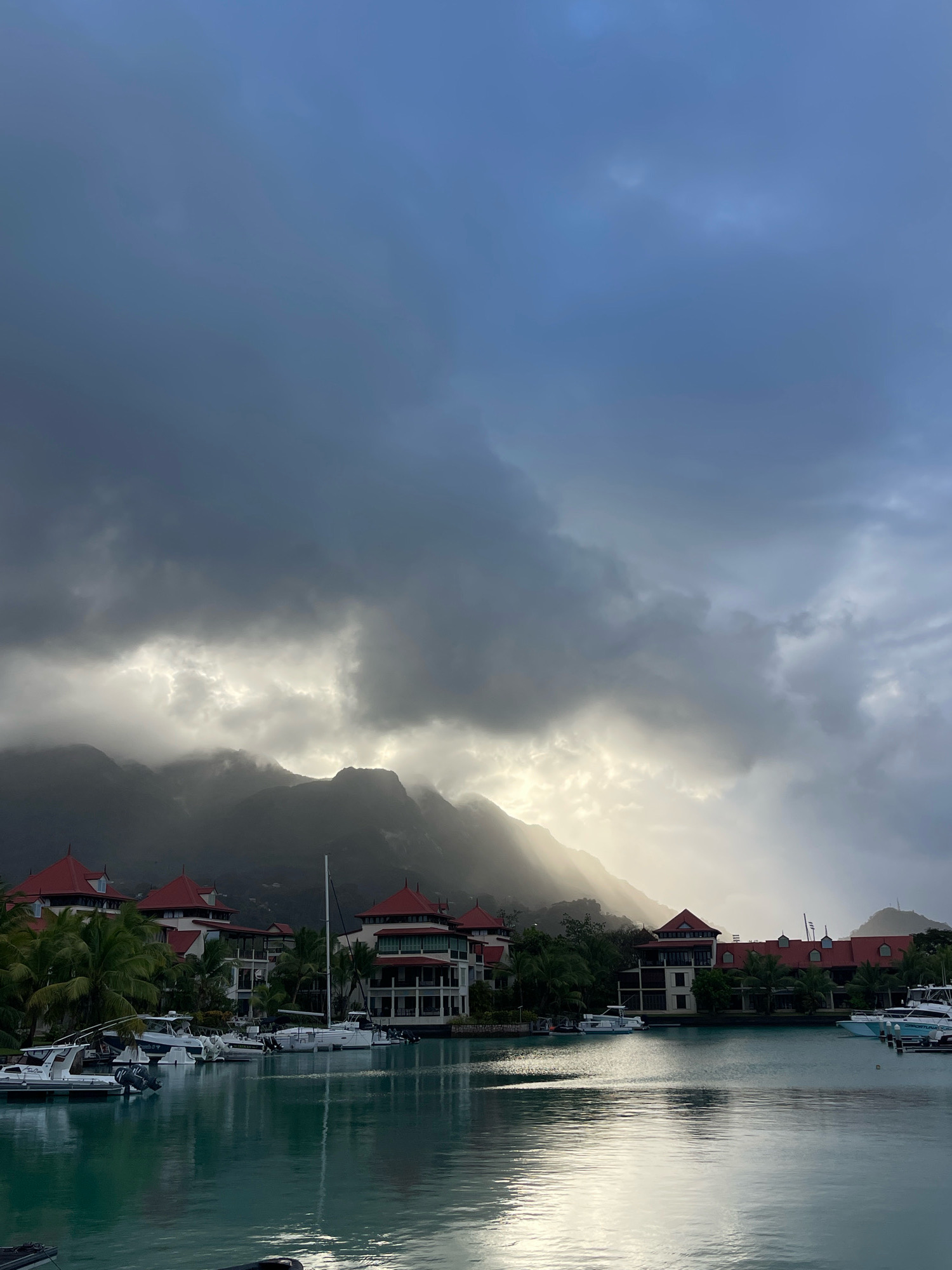 The image depicts a harbor in Providence, Seychelles, under a dramatic sky. In the foreground, calm, turquoise water reflects the light. Moored boats of various sizes are lined up along the waterfront. Behind the boats, a row of buildings with distinctive red tiled roofs and white facades are visible, interspersed with palm trees. In the background, dark, imposing mountains are shrouded in clouds, with shafts of sunlight breaking through, illuminating the upper slopes. The overall scene conveys a sense of moody, tropical beauty.