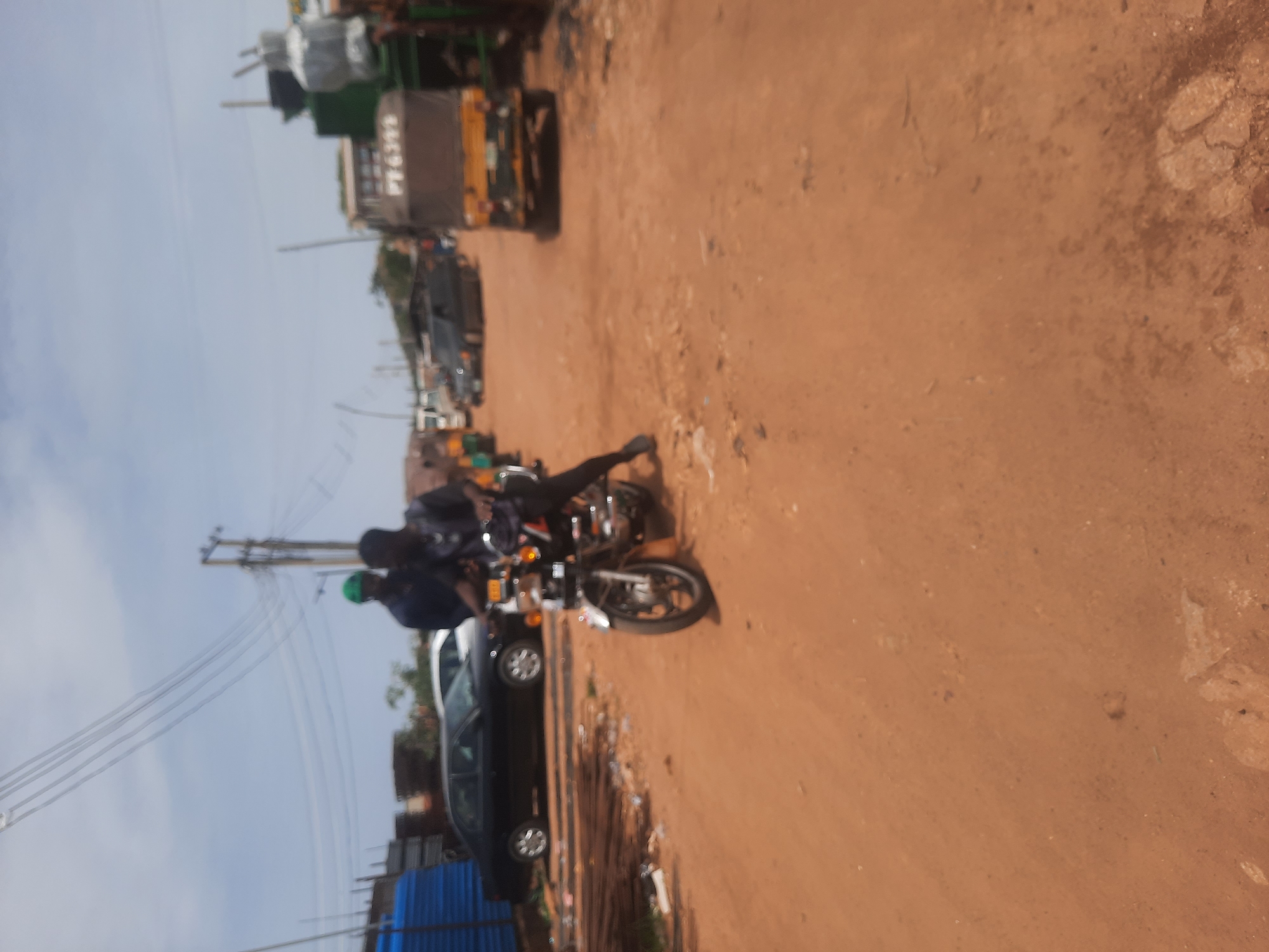 The image depicts a dirt road in Gura Top, Nigeria, with various vehicles parked along the roadside. A motorcycle with two riders is positioned near a black car. Further down the road, a collection of commercial vehicles or trucks are visible. A series of utility poles and overhead wires are present. The environment appears to be a typical street scene in a developing urban area.