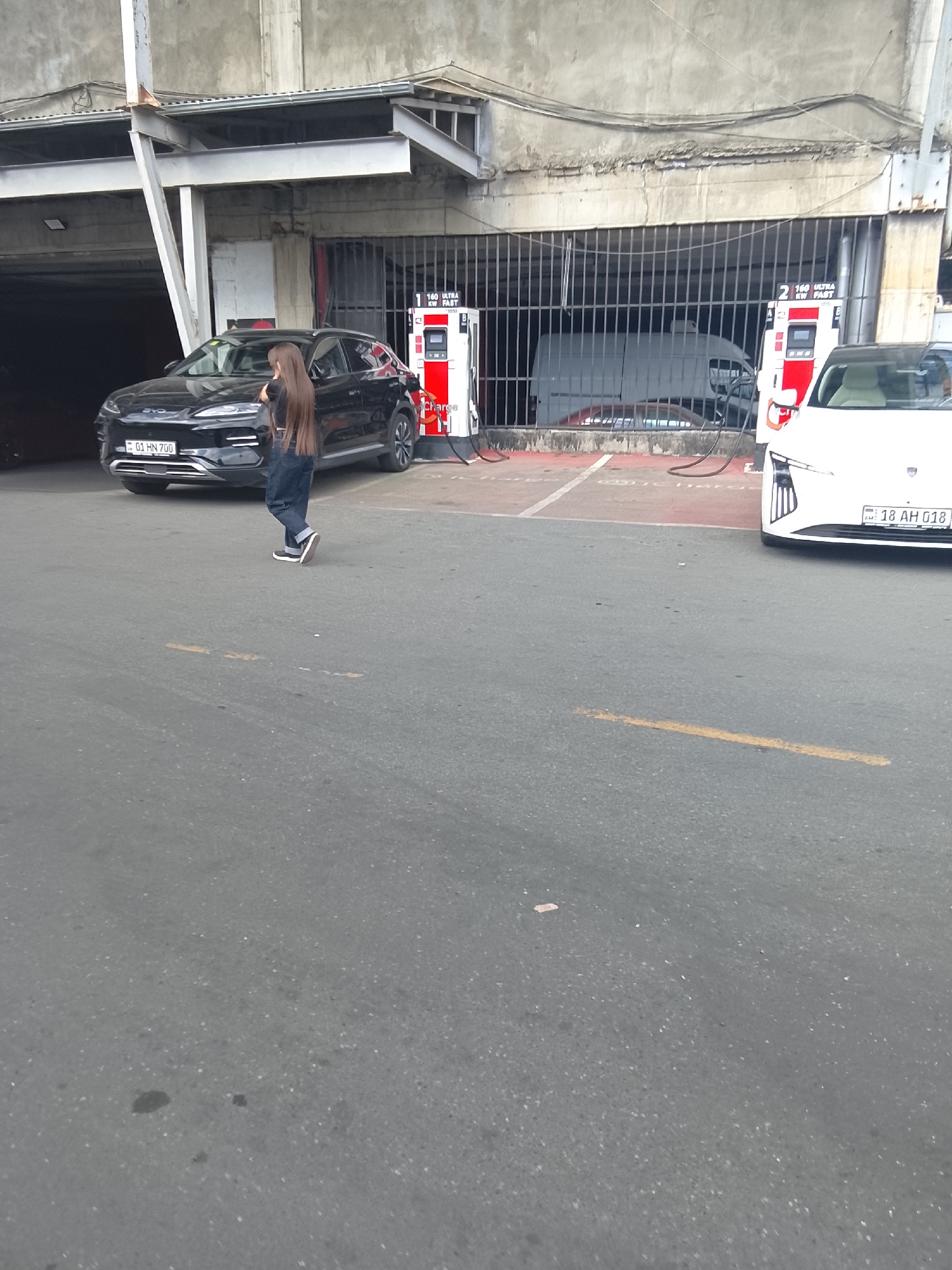 The image captures an outdoor charging station next to a concrete building in Unknown, Unknown. In the foreground, a person with long, straight, dark brown hair, wearing a dark top, dark blue jeans, and dark sneakers, is walking from left to right across a gray asphalt surface. Behind the person, a black electric BYD SUV with license plate "01 HN 700" is parked and connected to a red and white "160 ULTRA KW FAST" charging station, labeled "1". To the right, a white electric sedan with license plate "18 AH 018" is parked and connected to another identical charging station, labeled "2". A white cargo van is partially visible behind a metal grate barrier within an opening in the concrete building. The building itself is a multi-level concrete structure with visible wear, featuring an overhang and structural beams. Red markings are visible on the asphalt in the charging bays, and a yellow line is painted on the asphalt in the middle-right foreground.
