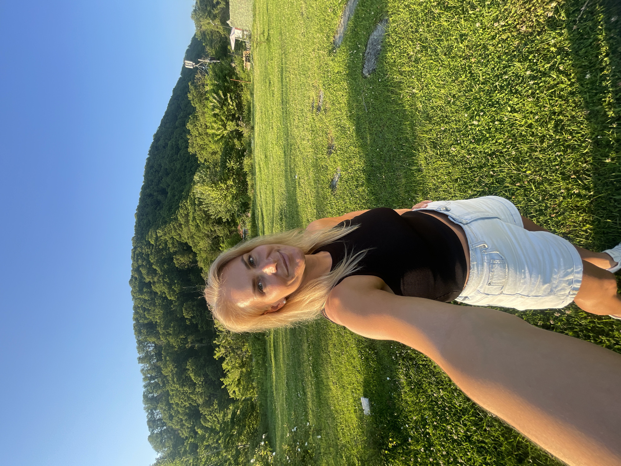 The scene is set outdoors in the city of Sochi, Russia, depicting a blonde female captured in a selfie. She is smiling and looking directly at the camera, holding the camera with her right arm extended towards the viewer. She is wearing a black ribbed sleeveless crop top, light blue denim shorts, and partially visible white sneakers. A small white stud earring is visible in her right ear.
The environment consists of a vibrant green grassy field in the foreground, interspersed with small white flowers, possibly clover. Several dark, irregularly shaped stones or stepping stones are embedded in the grass. Long shadows stretch across the ground, indicative of a low sun angle, suggesting either late afternoon or early morning.
In the mid-ground and background, dense green trees cover a large hill or mountain that rises under a clear, bright blue sky. On the right side of the hill, a prominent cellular communication tower, a metal trellis structure with multiple antennas, is visible amidst the trees. Further in the far right background, a white umbrella or tent-like structure is present, along with other indistinct structures. A small, indistinct building or structure is partially visible through the trees in the far left background.