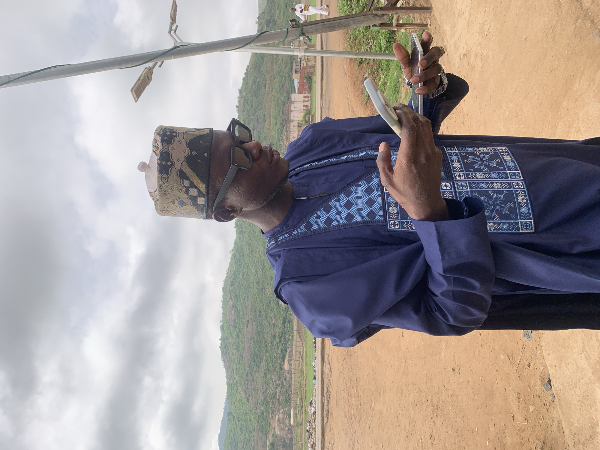 The image shows a man in Jos, Nigeria. He is wearing a blue traditional garment with intricate embroidery and a patterned cap. He also wears sunglasses and a watch. He holds a white smartphone in his hands, appearing to be focused on it.
The background features a landscape with hills and a cloudy sky. There is a dirt ground area. A light post is visible in the frame. In the distance, there appears to be another person wearing white clothing. The environment suggests an outdoor setting in Jos.