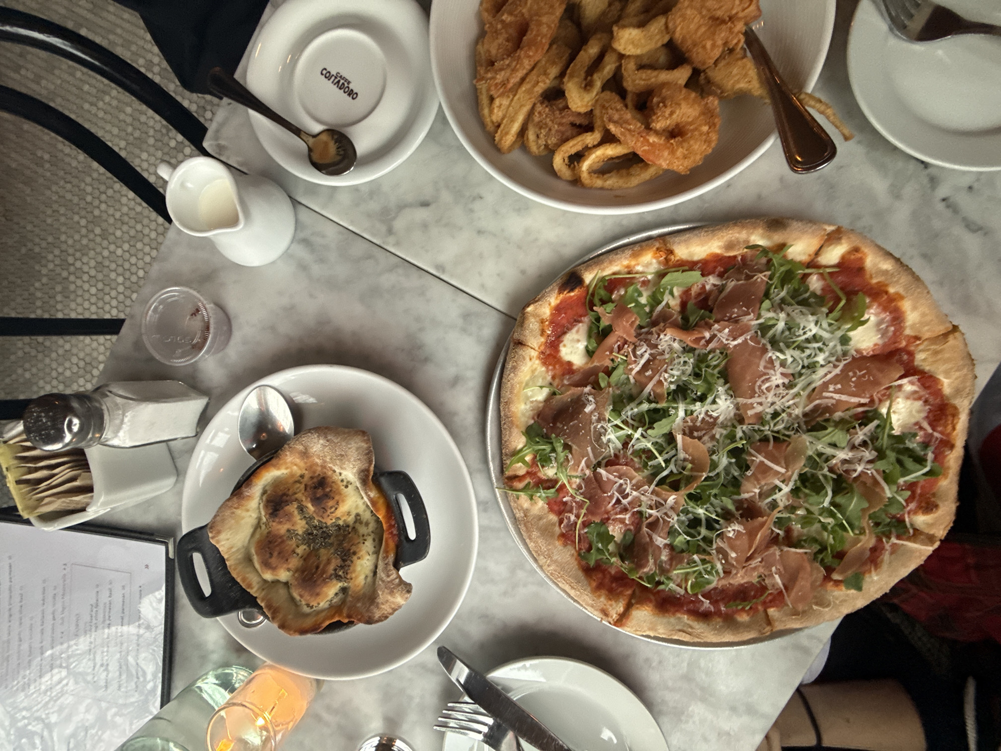 A high-angle shot depicts a table at a restaurant in New York City. The table is covered in a light marble surface. On the table, there is a pizza topped with arugula and prosciutto, a bowl of fried calamari, and a dish with a bread-covered top, all on white plates. There are also several smaller dishes, silverware, a creamer, a salt and pepper shaker, a menu, and a lit candle. The restaurant has a black-and-white tiled floor.