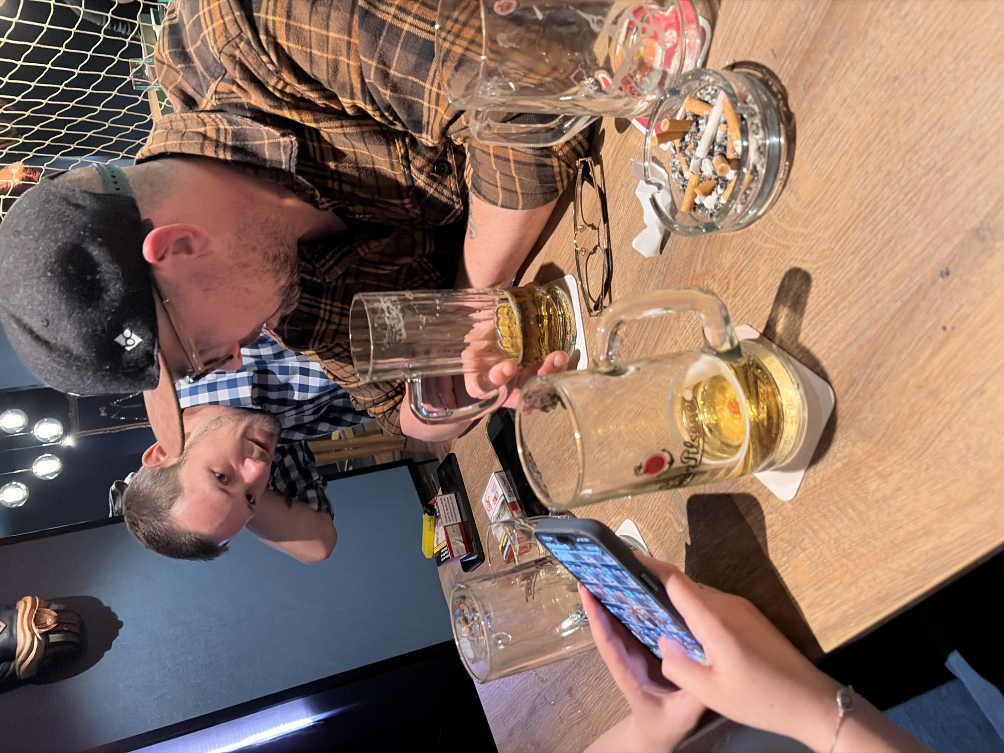 The image depicts an indoor scene, likely a bar or restaurant, with a wooden table as the primary surface. Three individuals are visible. In the foreground, a person's hands are holding a smartphone, displaying colorful images on its screen. To the left, a glass mug filled with a golden liquid, presumably beer, sits on a coaster. Next to it, another glass mug, empty, is positioned. Further to the right, an ashtray contains several cigarette butts and ash. Two other individuals are partially visible in the background. One man, wearing a plaid shirt, is leaning forward, looking down. Another man, also in a plaid shirt, has his back to the camera, wearing a black baseball cap. The lighting suggests an evening setting. The presence of a fishing net in the background and the mention of Langeoog, Germany, suggest the location might be a coastal establishment on the island of Langeoog.