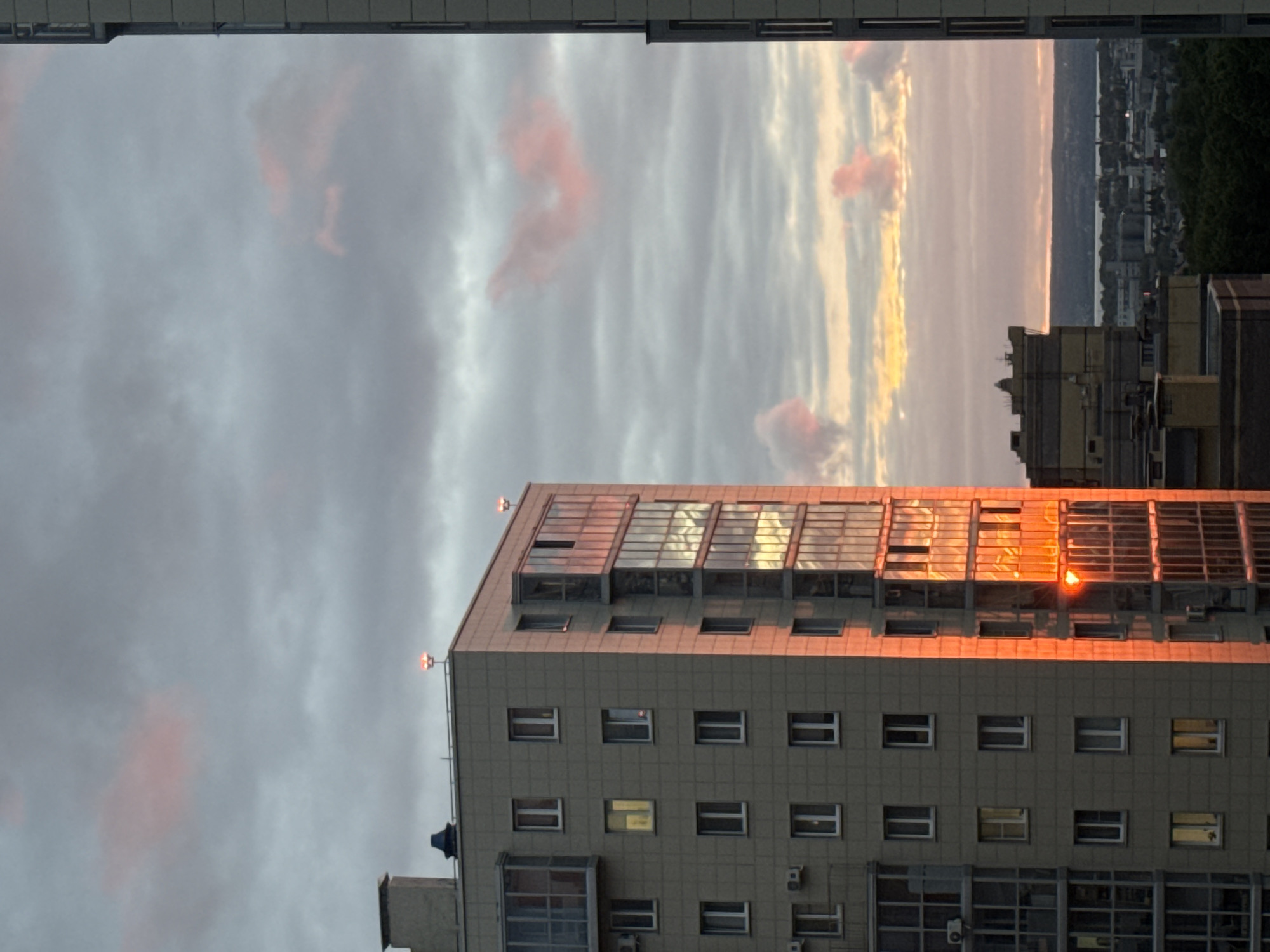 The image depicts a tall, gray building in Kazan, Russia, illuminated by the golden light of a setting sun. The building has multiple windows and a glazed upper section that reflects the sunlight intensely. Above the building is a cloudy sky with pink-tinged clouds, suggesting the time of day. In the distance, other buildings and possibly a body of water can be seen. The image is taken from an elevated position, looking slightly upward at the building.