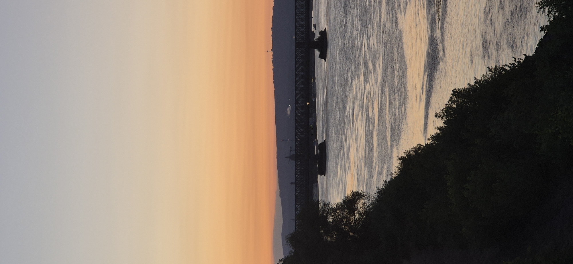 This image depicts a sunset over the Rhine River in Mainz, Germany. The sky transitions from a pale blue at the top to a warm orange and yellow gradient towards the horizon. A silhouetted bridge, likely the Theodor Heuss Bridge, spans the river in the midground, supported by pillars rising from the water. Numerous boats are visible on the river. On the far bank, a low-lying silhouette of hills and buildings is present, with a prominent radio tower visible on the highest point. In the foreground, a dark, silhouetted hillside covered in dense foliage slopes down towards the river. The water's surface reflects the colors of the sunset, creating shimmering patterns.