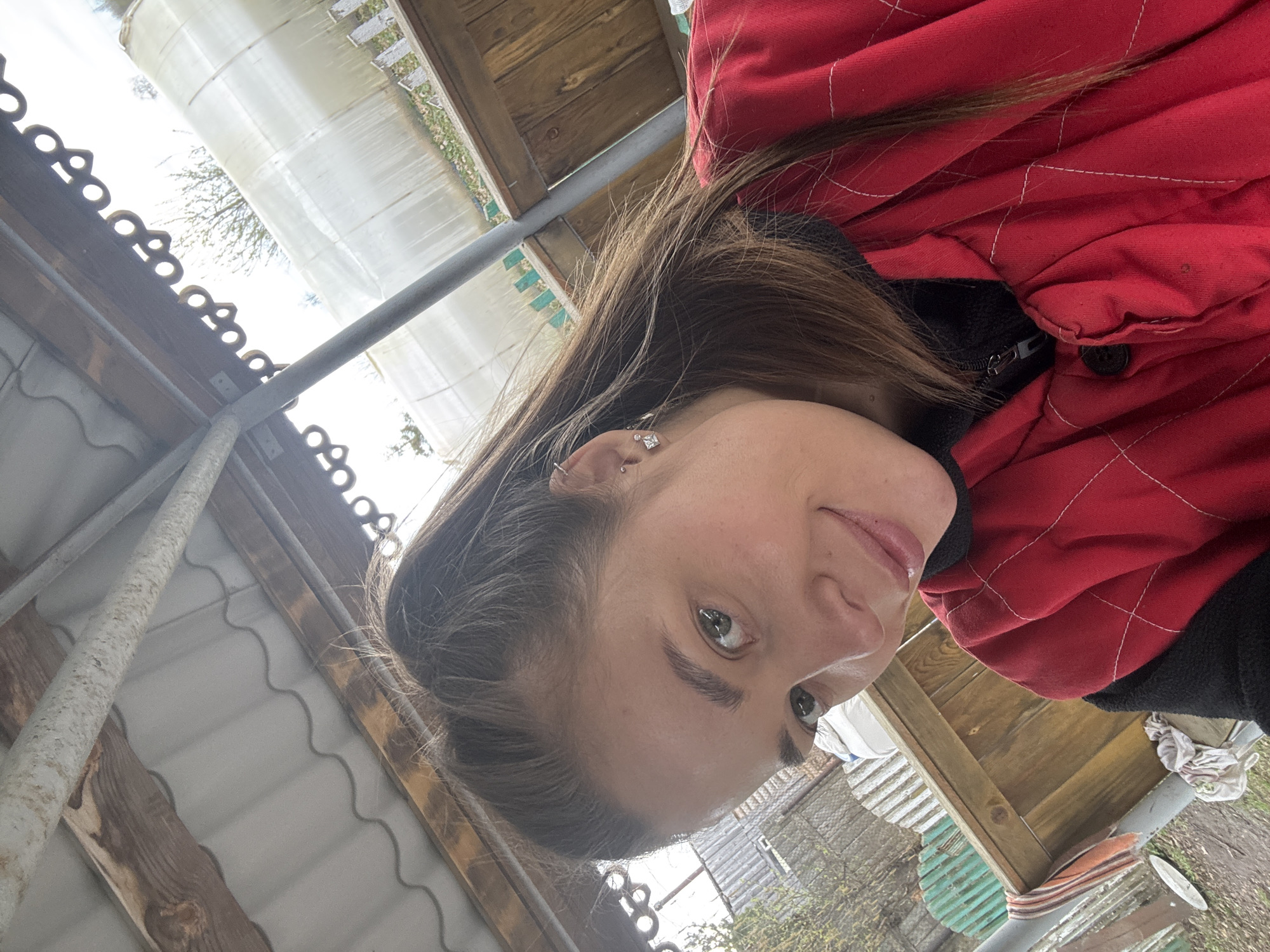 This image is a portrait-style photograph of a woman taken outdoors, likely a selfie, given the low and slightly skewed angle. The woman, appearing to be in her 20s or 30s, is the primary subject, filling much of the frame with her head and upper torso. She has long, dark brown hair, green eyes, and a subtle, pleasant expression, with a slight smile or smirk. She is wearing a red quilted jacket with visible white stitching, layered over a black zippered garment. In her visible ear, she has two earrings: a small stud and a larger, decorative square-shaped dangle.
The background reveals an outdoor, utilitarian setting, consistent with a rural or backyard area in Сульмаш, Russia. Above and behind the woman, various structures are visible, including wooden planks, possibly forming part of a shed or roof, and sections of corrugated metal or plastic sheeting. Metal poles or pipes run vertically and diagonally, suggesting structural supports. In the further background, a large expanse of translucent plastic sheeting, indicative of a greenhouse or a covered outdoor structure, is prominent. Through this sheeting and above the structures, bare tree branches can be seen, hinting at either early spring or late autumn. The lighting appears soft and diffused, typical of an overcast day, without harsh shadows. A crumpled white and orange striped cloth and another white cloth are visible on a wooden surface near a metal pipe in the bottom right corner. There is no visible text in the image.