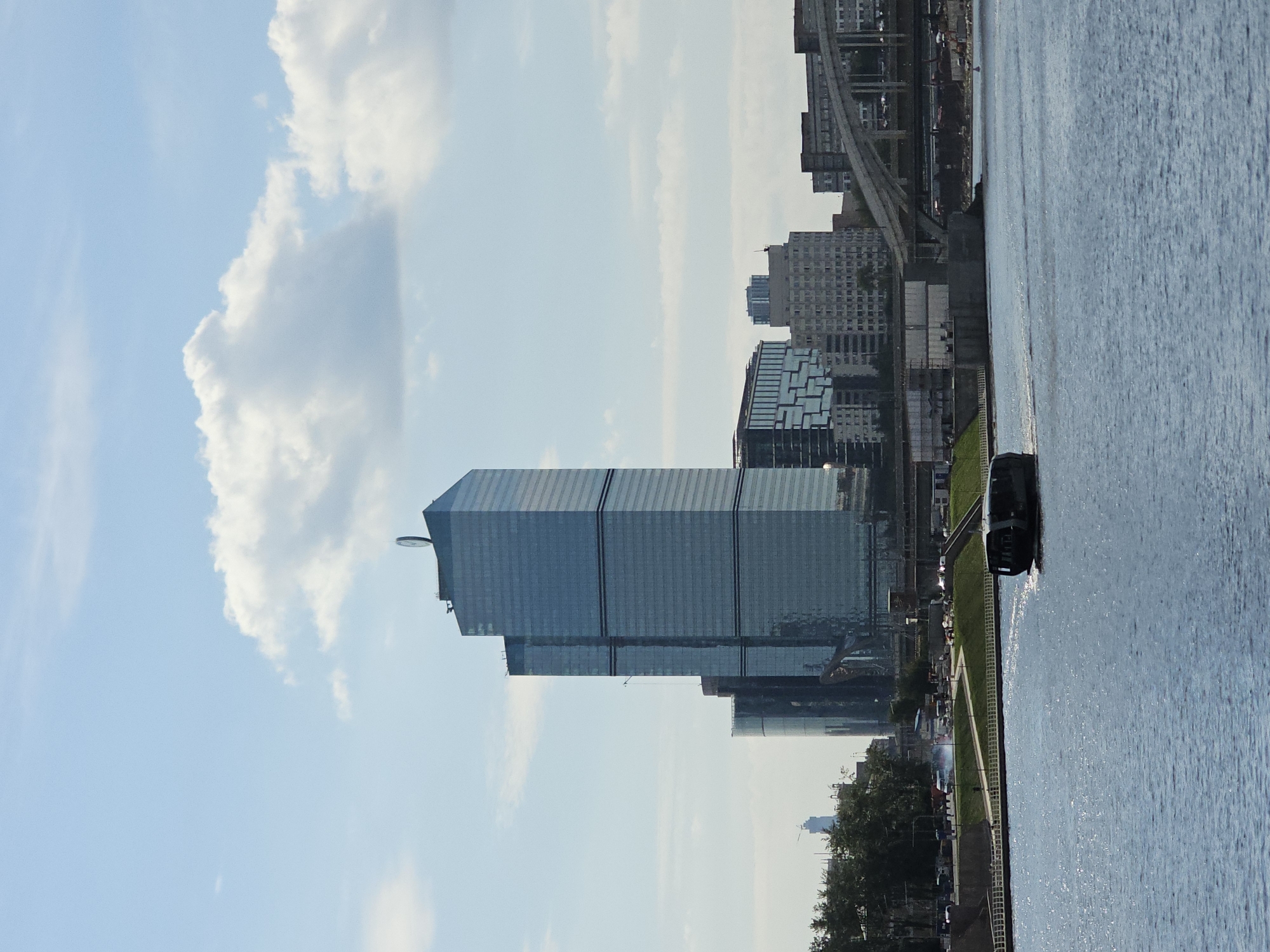 This is a daytime, eye-level shot of a wide, blue body of water in the foreground, reflecting the bright sky and passing clouds. A dark, sleek boat with a curved hull travels across the water from left to right. In the middle ground, a grassy embankment with a white railing separates the water from a busy urban scene. A road with several vehicles is visible behind the embankment, and a cluster of modern glass skyscrapers dominates the background. The tallest building, a slender, rectangular skyscraper with dark glass and horizontal banding, has a circular emblem at its peak. To the right of the skyscrapers, an arched bridge spans a section of the urban landscape. The sky is a clear blue with scattered white clouds. The scene appears to be a waterfront view of the Moscow International Business Center, also known as Moscow City, a business district in western Moscow, Russia.