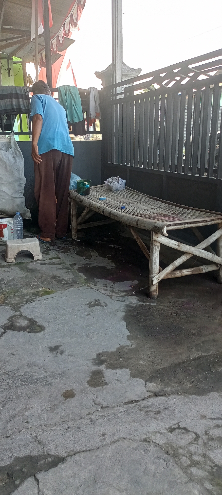 In Gempolklutuk, Indonesia, a person wearing a blue shirt and brown pants stands next to a large white sack. In front of them, a small white stool holds a plastic water bottle. To the right, a low bamboo bed is situated on a concrete floor that appears wet and stained. A green bucket and a clear plastic container with white fabric are on the bed. A dark metal fence is visible in the background, with clothing hanging on a line above it. The scene is outdoors, under what appears to be a covered structure.