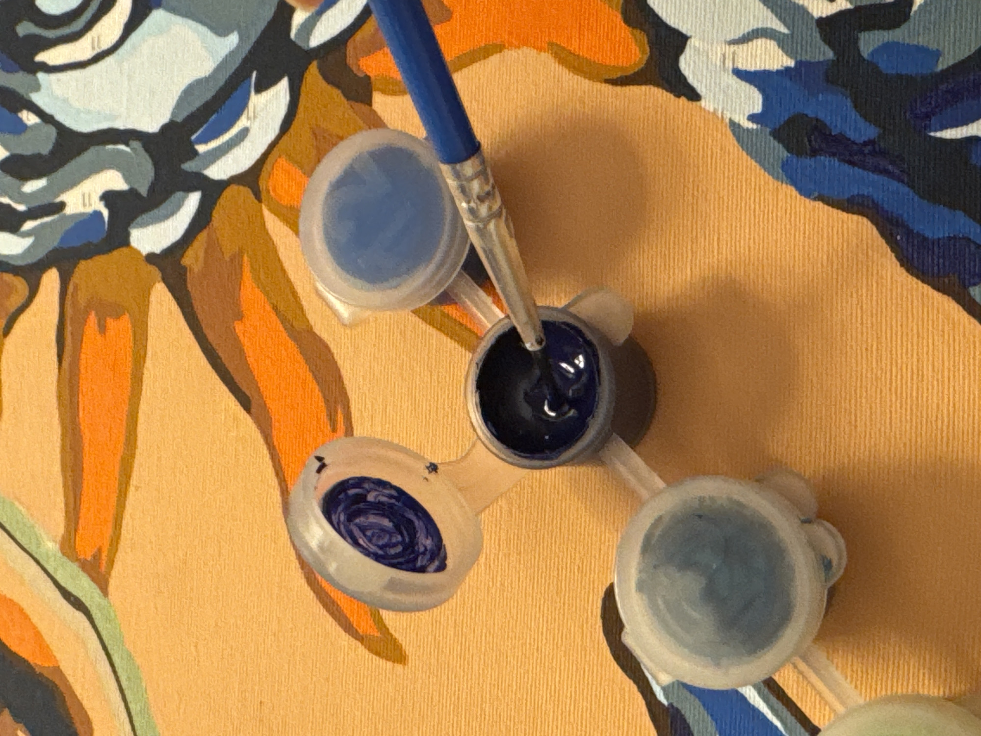 The image shows a close-up of a paint-by-numbers artwork in progress. There are several small plastic containers with paint lids, each holding a different color of paint. A paintbrush with a blue handle is dipping into a container of dark blue paint. The background of the artwork is a textured orange surface, and parts of the painting show areas of orange, blue, and white. There is no indication of the city of Aksu, Turkey, or any specific location in the image.
