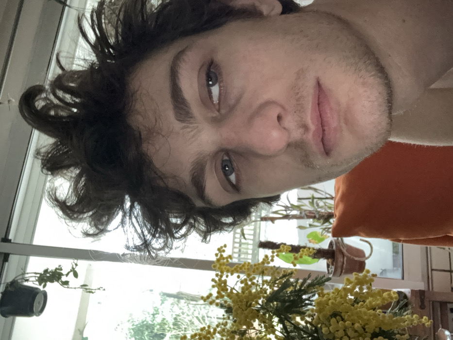 The image displays an adult male within an indoor setting in Vincennes, France. The subject, a Caucasian male, occupies the majority of the frame with dark, disheveled curly hair, light-colored eyes, and light stubble on his chin and upper lip. He looks directly forward, with his visible left shoulder appearing bare. An orange cushion is partially visible behind his right shoulder. To the left of the subject, a significant arrangement of bright yellow mimosa flowers is prominently displayed. The background shows a large window revealing an outdoor scene, which includes a white railing, various potted plants, a hanging plant, and distant green foliage.