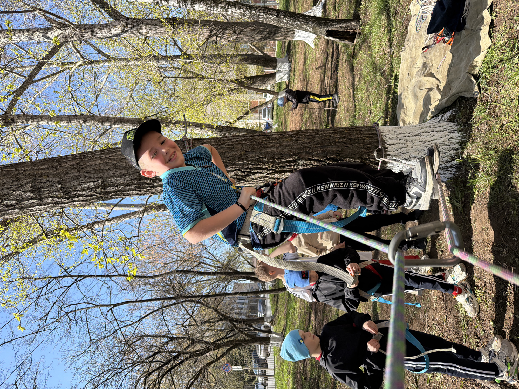A group of children are participating in a high ropes course or similar outdoor adventure activity in a park-like setting. The foreground focuses on one boy wearing a blue and green striped shirt, black pants with white stripes, and a black cap, who is suspended between trees using a harness and ropes. He is smiling and appears to be enjoying the activity. Other children, also wearing harnesses and connected to ropes, are visible behind him, partially obscured by the ropes and the tree trunk. The environment consists of trees with budding leaves, grass, and other park vegetation. In the background, there is a building and a signpost visible. The location is identified as Izhevsk, Russia, based on contextual information.