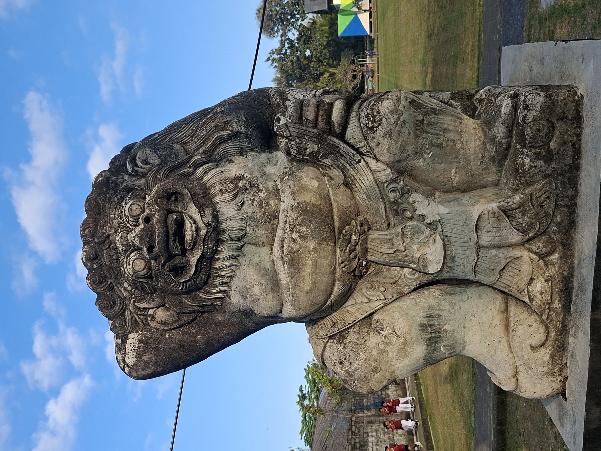 An imposing, weathered stone statue of a mythical creature, possibly a Rakshasa or a guardian spirit, stands prominently in the foreground against a bright blue sky with scattered white clouds. The statue is intricately carved, depicting a fierce face with wide eyes, an open mouth revealing teeth, and a mane of swirling hair. It has a large, rotund body with elaborate clothing or armor. The creature's left arm is raised, resting on its head, while its right arm appears to be holding something. The statue sits on a concrete base.
In the background, to the left of the statue, a group of children in red shirts and white pants are walking on a paved path. Behind them is a low, gray brick wall and some green trees. To the right of the statue, a grassy field stretches out, with more trees and a building with colorful geometric panels. A thin black wire runs horizontally across the upper portion of the image, passing in front of the sky. The overall impression is of an outdoor setting in a place with a cultural heritage, possibly a temple or a public park in Blahbatuh, Indonesia. The lighting suggests it is daytime, with the sun casting subtle shadows.