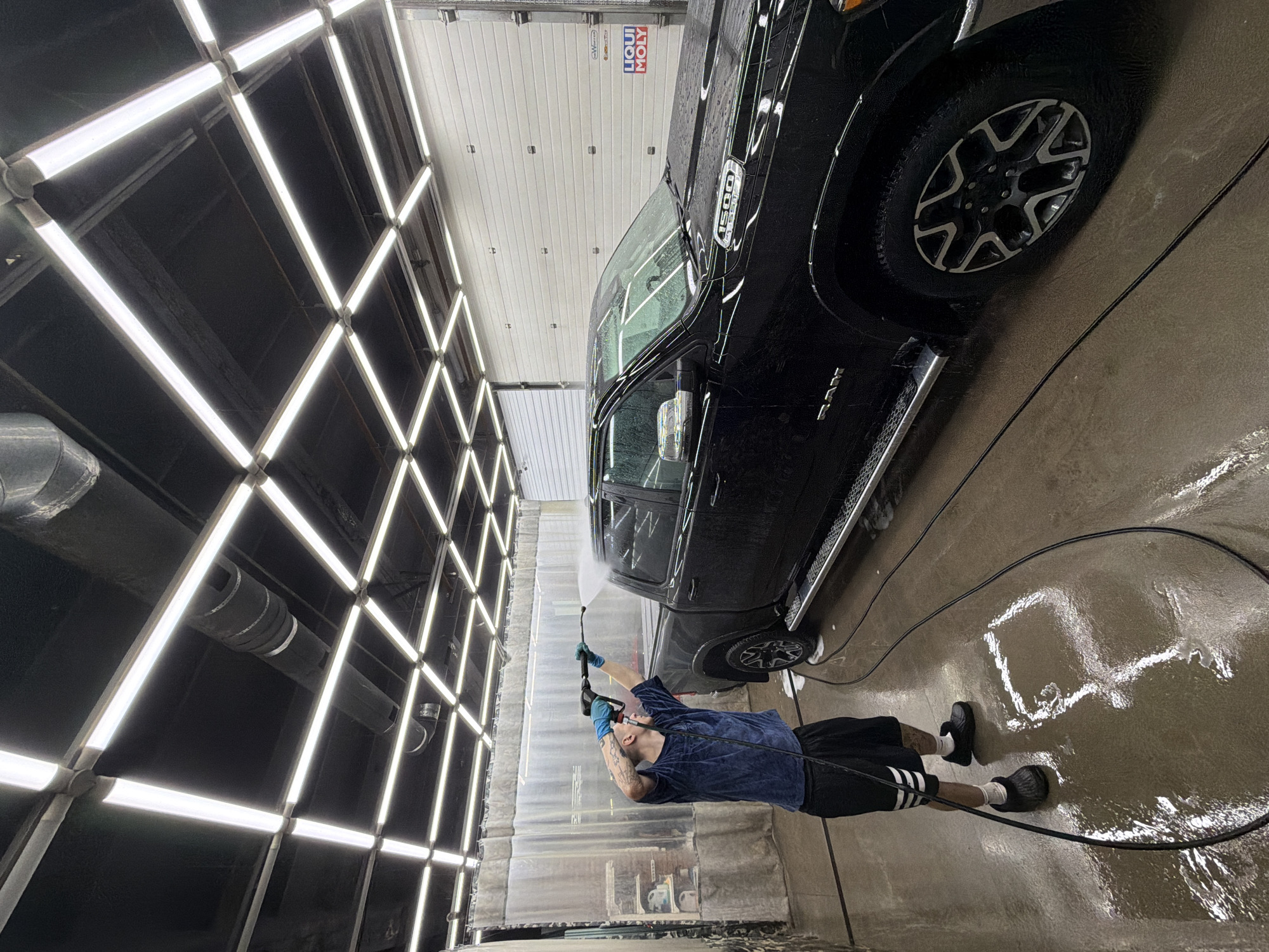 A man in a blue shirt and black shorts is pressure-washing a black Ram 1500 pickup truck in what appears to be an automated car wash facility. The floor is wet, suggesting recent or ongoing washing. The walls and ceiling are lined with bright fluorescent lights arranged in a grid pattern. A sign for Liqui Moly is visible on the back wall of the car wash. The location is identified by the car wash brand as being in Krasnoyarsk, Russia.