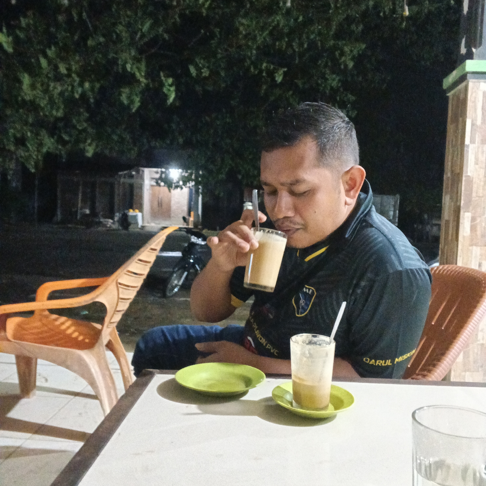 At night, a man is seated at a white table, drinking from a clear glass of iced coffee with a straw. He is wearing a dark short-sleeved shirt with yellow trim and a logo on the left chest. To his right, another glass of iced coffee sits on a small green saucer. In the background, an orange plastic chair is visible, and beyond that, trees and a building with a pitched roof. A motorcycle is parked to the left of the orange chair. The scene appears to be an outdoor seating area.