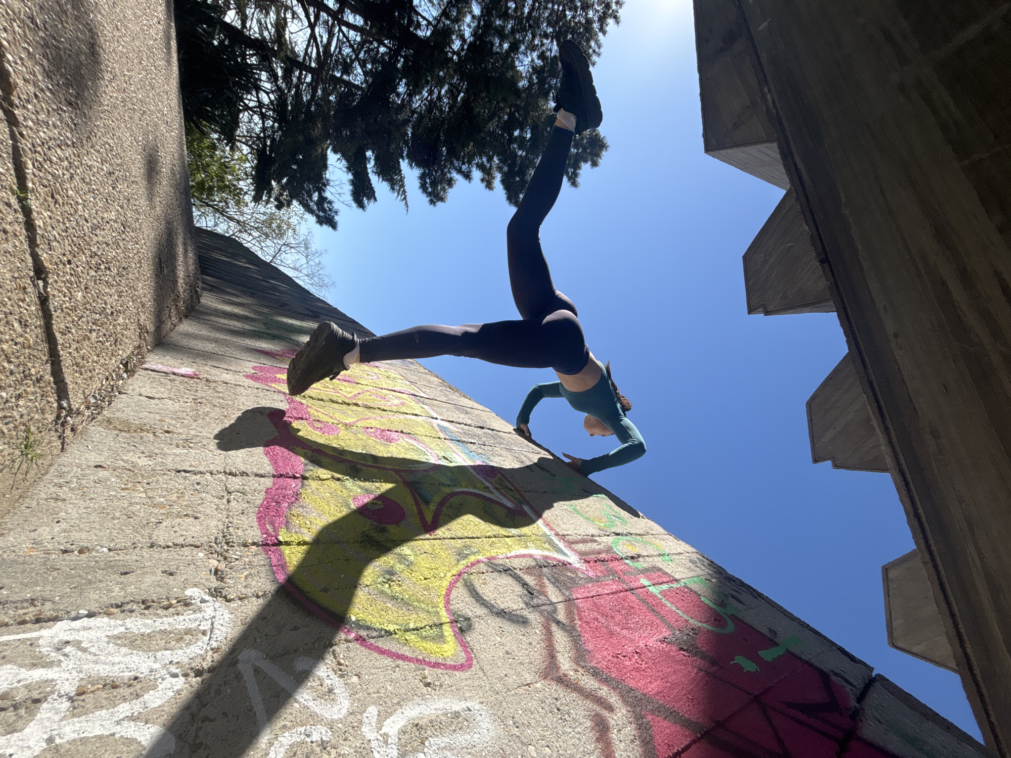 An acrobatic shot captures a person performing a dynamic inverted pose against a sloped, graffiti-covered concrete wall under a clear blue sky. The image is taken from a low angle, looking up, with the subject positioned diagonally across the frame.
The central subject is a person, likely a young adult female, wearing dark athletic leggings and a long-sleeved teal top. They are in a handstand or a similar balancing pose, with their hands placed on the textured concrete surface and their body extended upwards and slightly outwards. One leg is bent at the knee, with the foot near the concrete, while the other leg is extended straight, reaching towards the top right of the frame. The word "VOOA" is visible on the left thigh of their leggings.
The setting appears to be an outdoor urban or industrial space, characterized by robust concrete structures. The sloped surface the person is on is heavily adorned with bright graffiti in shades of yellow, pink, and green, interspersed with some indistinct white and black markings. To the right, angular concrete architectural elements are visible, framing a portion of the bright blue sky. Above and behind the person, dark green foliage from trees is silhouetted against the bright sunlight.
The strong, crisp shadows cast by the person and the surrounding structures indicate bright sunshine, suggesting it is daytime, possibly late morning or early afternoon. The overall impression is one of athleticism, balance, and creative use of an urban environment.