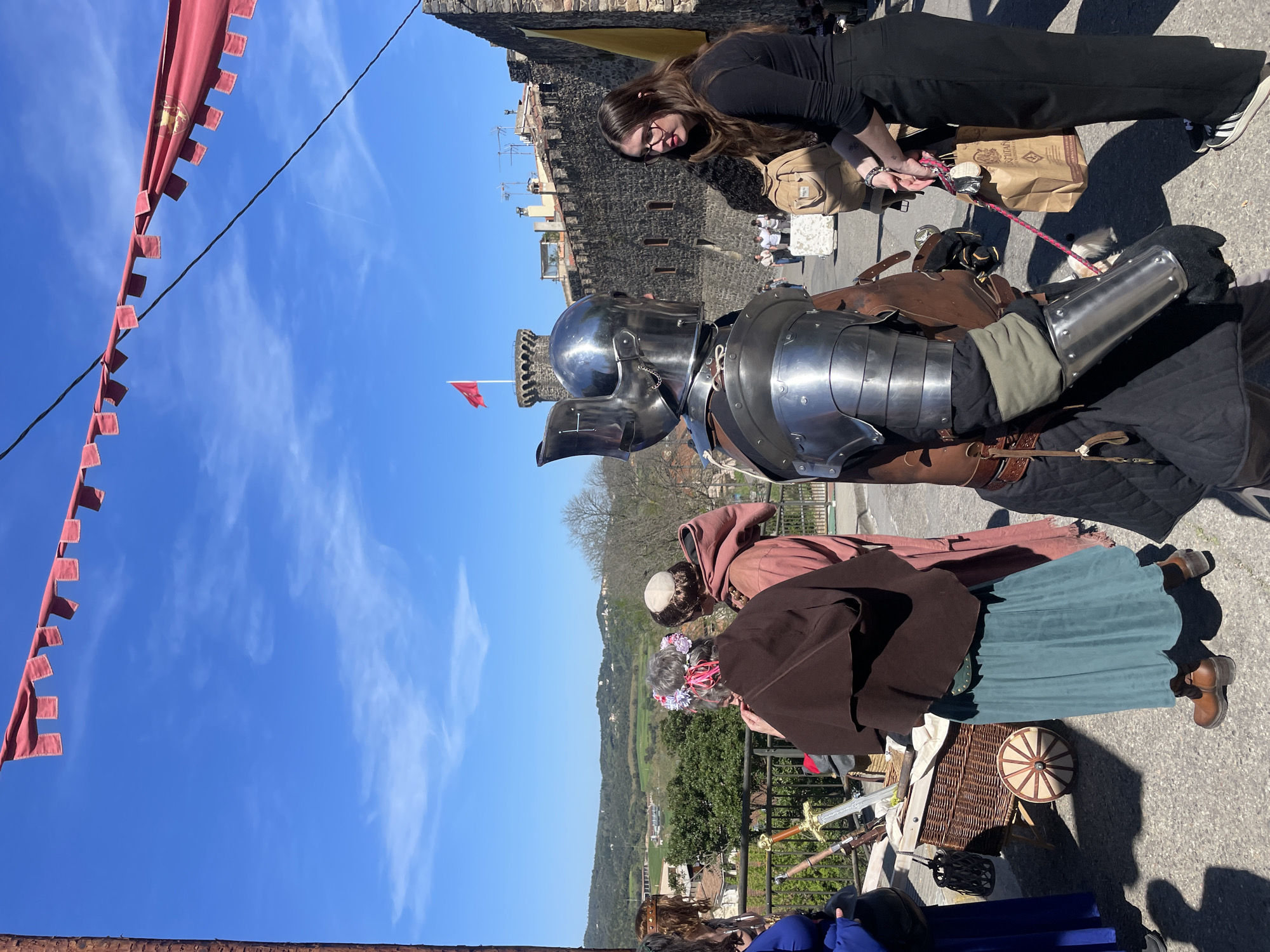 The image depicts a scene in Hostalric, Spain, characterized by a medieval reenactment or fair. A person dressed in full medieval armor stands prominently in the foreground, with a woman in black clothing adjusting something near the armor. Behind the armored figure are other individuals in medieval attire, including one in a brown robe, suggesting a monk or friar. A woman wearing a flower crown is visible, standing close to swords and a basket. In the background, the stone walls and towers of the Castle of Hostalric are visible. Red banners hang along the side, and the clear blue sky with scattered clouds serves as the backdrop. The landscape reveals buildings and green fields beyond the castle, which marks the general vicinity of Hostalric.