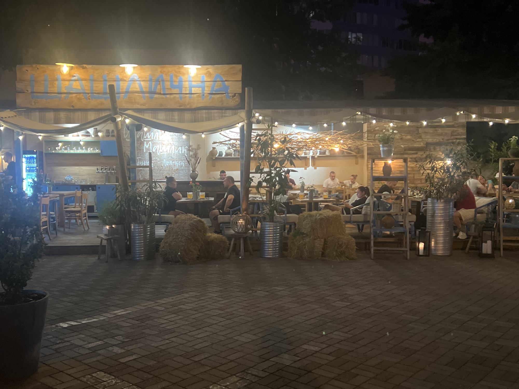 The image shows an outdoor seating area of a restaurant at night. The restaurant has a rustic, wooden facade and is decorated with string lights and straw bales. Several people are seated at tables, eating and drinking. Some people are standing or walking around. The ground is paved with bricks. In the background, a building with illuminated windows is visible. The overall atmosphere appears relaxed and convivial.