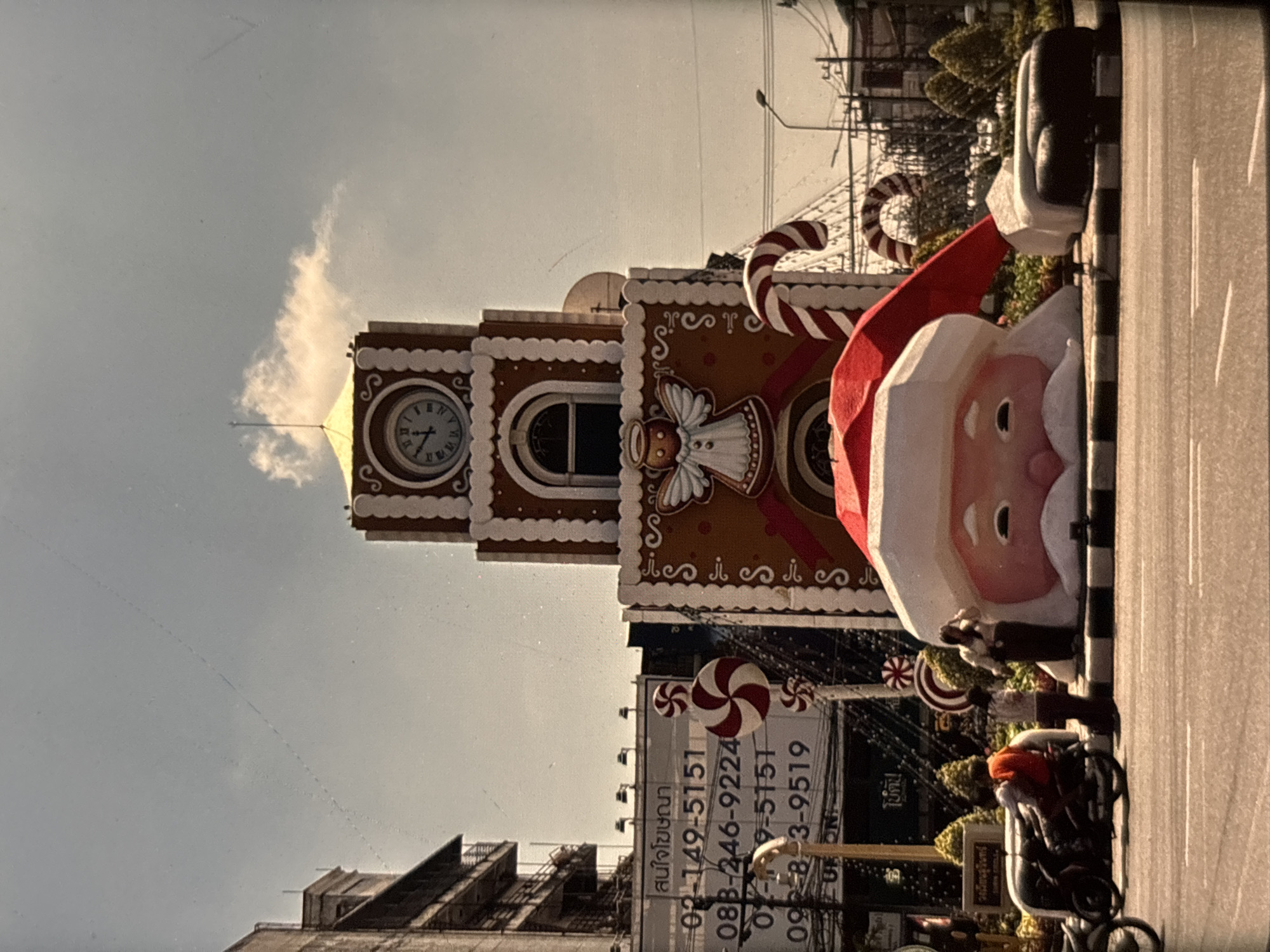 In Chalong, Thailand, a street scene unfolds under a bright sky featuring a large, gingerbread-themed clock tower decorated for Christmas. The tower displays a clock face with Roman numerals, and is adorned with white icing-like trim and windows. At the base of the tower, a giant Santa Claus head, with a red hat and white beard, rests on the ground. A large candy cane curves around the side of the gingerbread structure. In the foreground, a motorcycle is parked, and two people stand near the Santa head. One person is adjusting their clothing, while the other stands with their hands clasped. A billboard in the background displays Thai text and phone numbers. The street is paved, with white lines marking lanes. The overall setting suggests a festive display in a public area of the city.