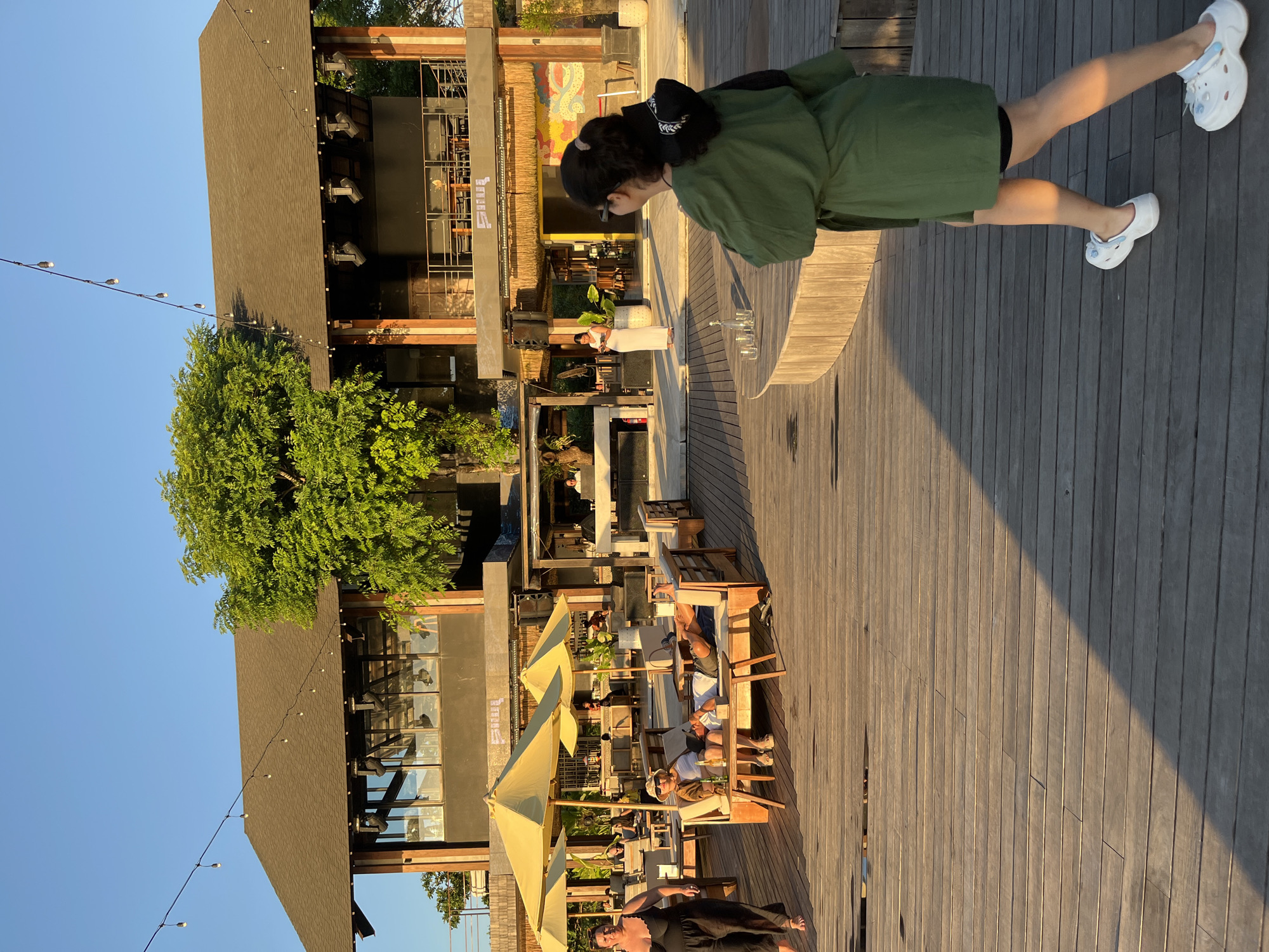 In Kuta Selatan, Indonesia, a woman in a green robe walks on a wooden deck towards a modern, multi-story building with a dark roof. The building has large windows and balconies, with lights and what appears to be a stage area. Several people are visible in the background, some seated on lounge furniture under yellow umbrellas, and others standing near the building. A tree with lush green foliage grows prominently in front of the building. The sun is setting, casting long shadows across the deck.