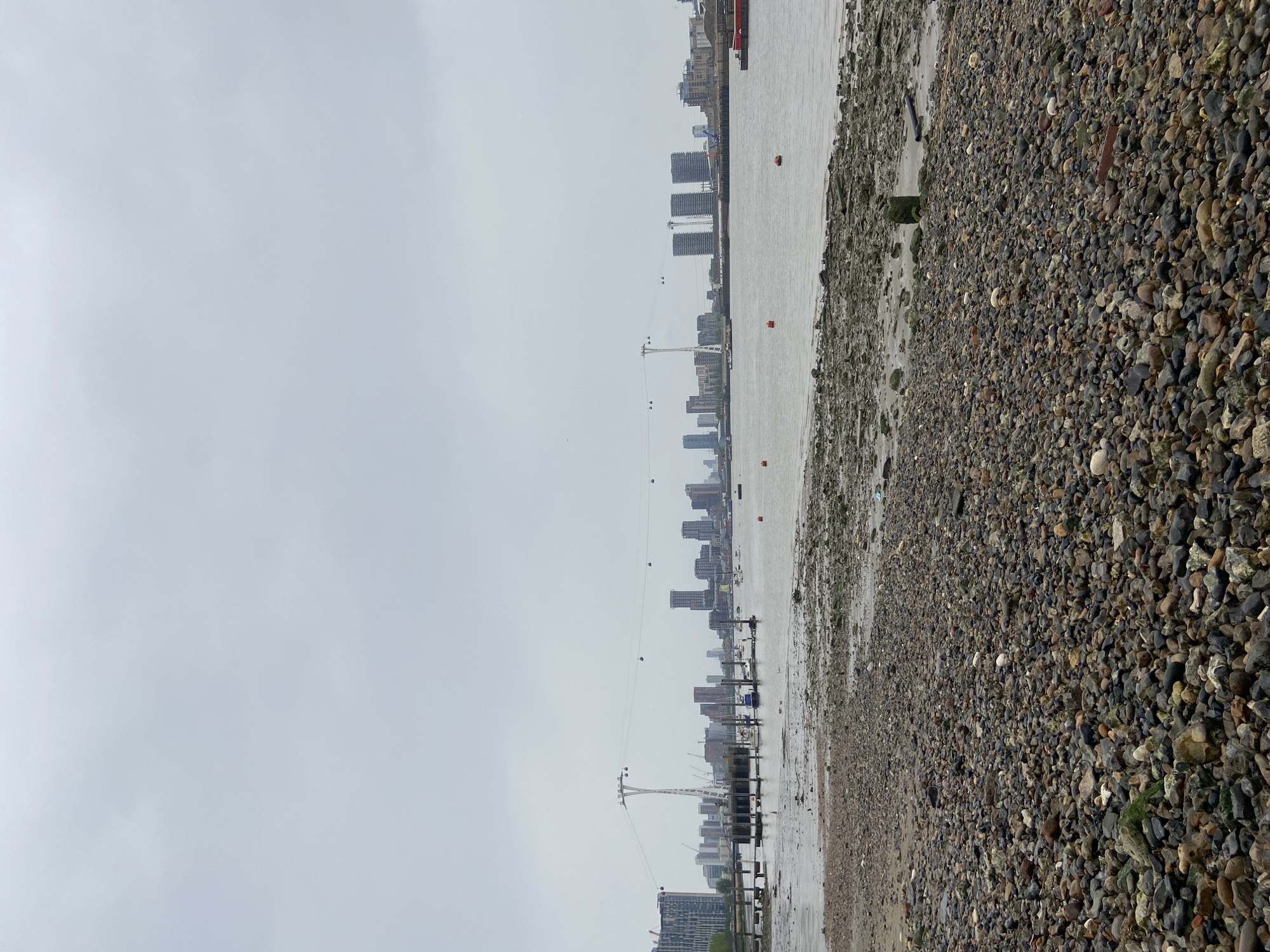 The image depicts a landscape view of the River Thames in London, United Kingdom. A pebble beach occupies the right foreground, transitioning to a muddy riverbank along the water's edge. The river flows horizontally across the center of the frame. The skyline of London is visible in the distance, punctuated by modern skyscrapers and the Emirates Air Line cable car. The sky is overcast and grey. No people are visible.