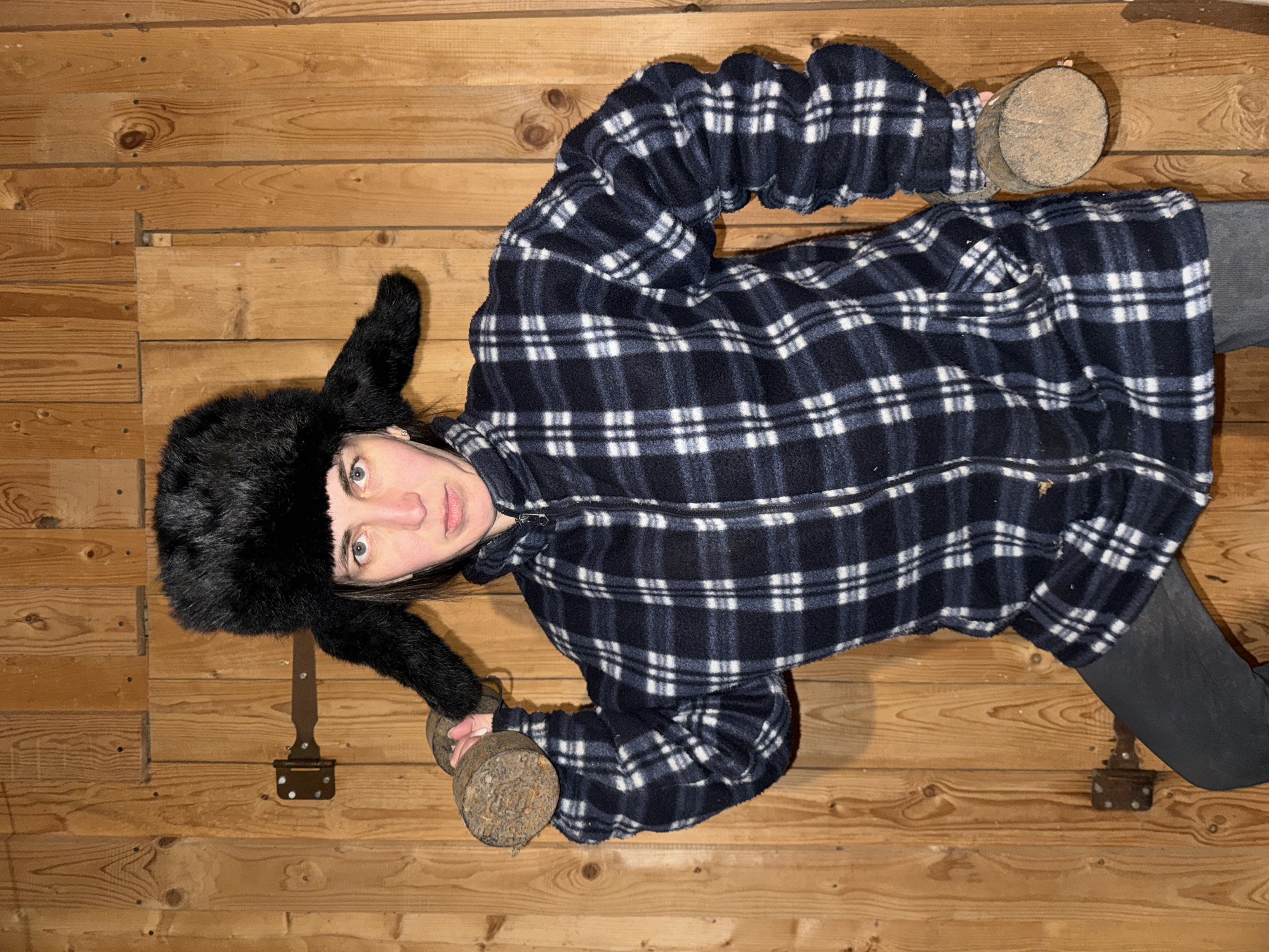 A single person is shown from an overhead perspective, appearing to lie on a wooden floor or stand against a wooden wall. The individual is wearing a black fur ushanka hat, a dark blue and white plaid fleece jacket with a zipper, and black leggings or trousers. They have fair skin, brown hair, and light-colored eyes, looking directly at the camera with a neutral to slightly serious expression.
The person is holding two rustic, cylindrical objects that resemble homemade dumbbells or weights. These objects appear to be made of a rough, earthy-colored material, possibly wood or concrete, with textured surfaces. One is held near their left shoulder, and the other near their right hip.
The setting is an indoor space with light brown wooden planks forming the entire background, showcasing visible grain, knots, and some fasteners. A dark metal latch or hinge mechanism is affixed to one of the planks behind the person's head.
The activity is a static pose, potentially for a humorous or unconventional photo related to exercise or strength, given the makeshift weights. The attire, particularly the ushanka, provides a cultural cue aligning with the location context of Вассята, Russia. No visible text is present. The time of day and weather are not discernible from the indoor scene.