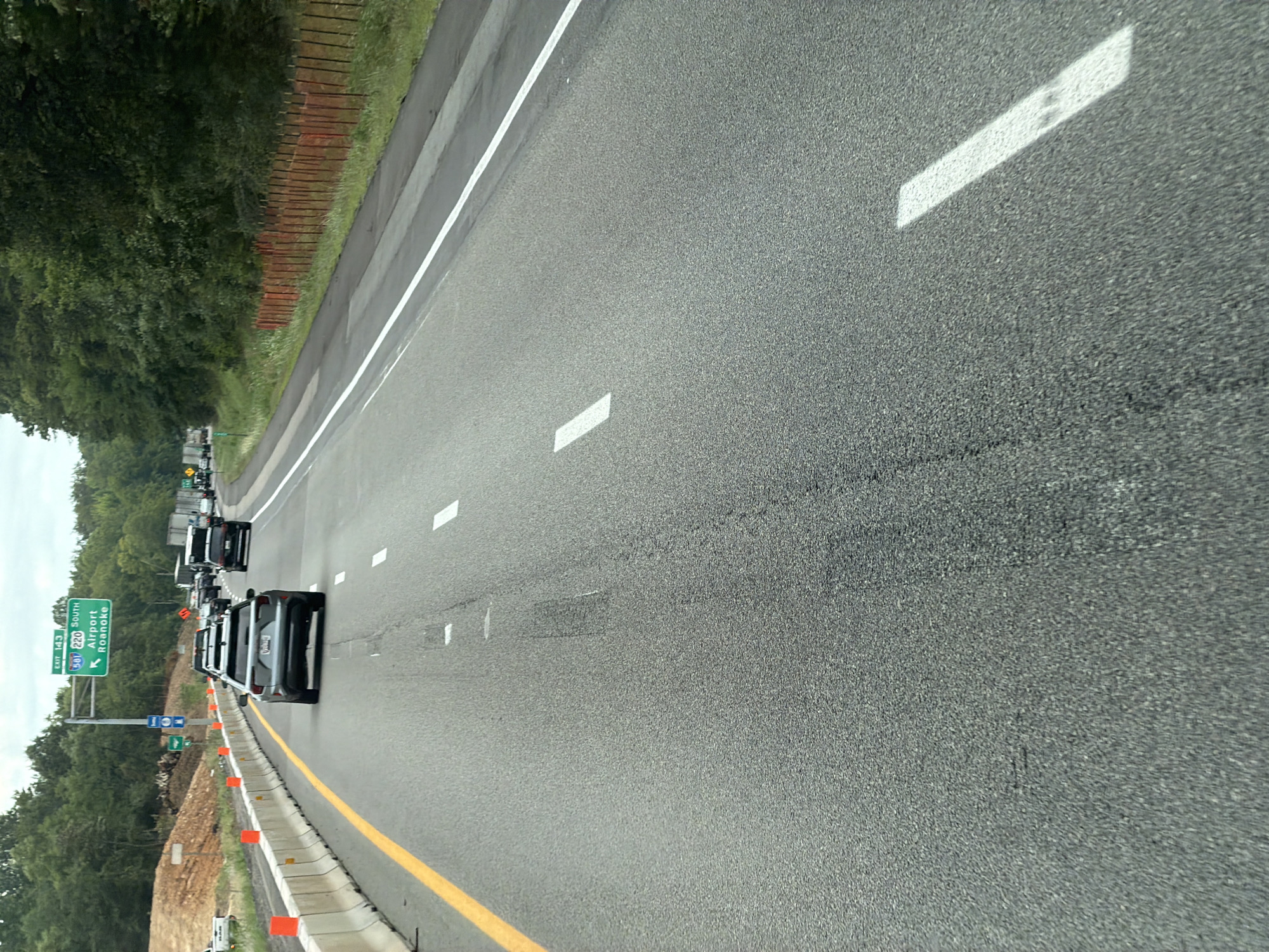 An eye-level, wide-angle exterior shot captures a highway scene in Hollins, Virginia, USA. The perspective is from a vehicle traveling on the right-hand lanes of a multi-lane highway. In the foreground, the asphalt road surface is visible with white lane markings. To the left, a yellow line separates the travel lanes from a construction zone, indicated by concrete barriers topped with orange cones. A few vehicles are on the highway, including a silver SUV in the right lane and several other cars and trucks further ahead. Overhead, a highway sign indicates "EXIT 143," "581 SOUTH 220 SOUTH," and points to "Airport" and "Roanoke." The background features a dense line of trees and vegetation on the right, with a construction site on the left with dirt mounds. The sky is overcast.