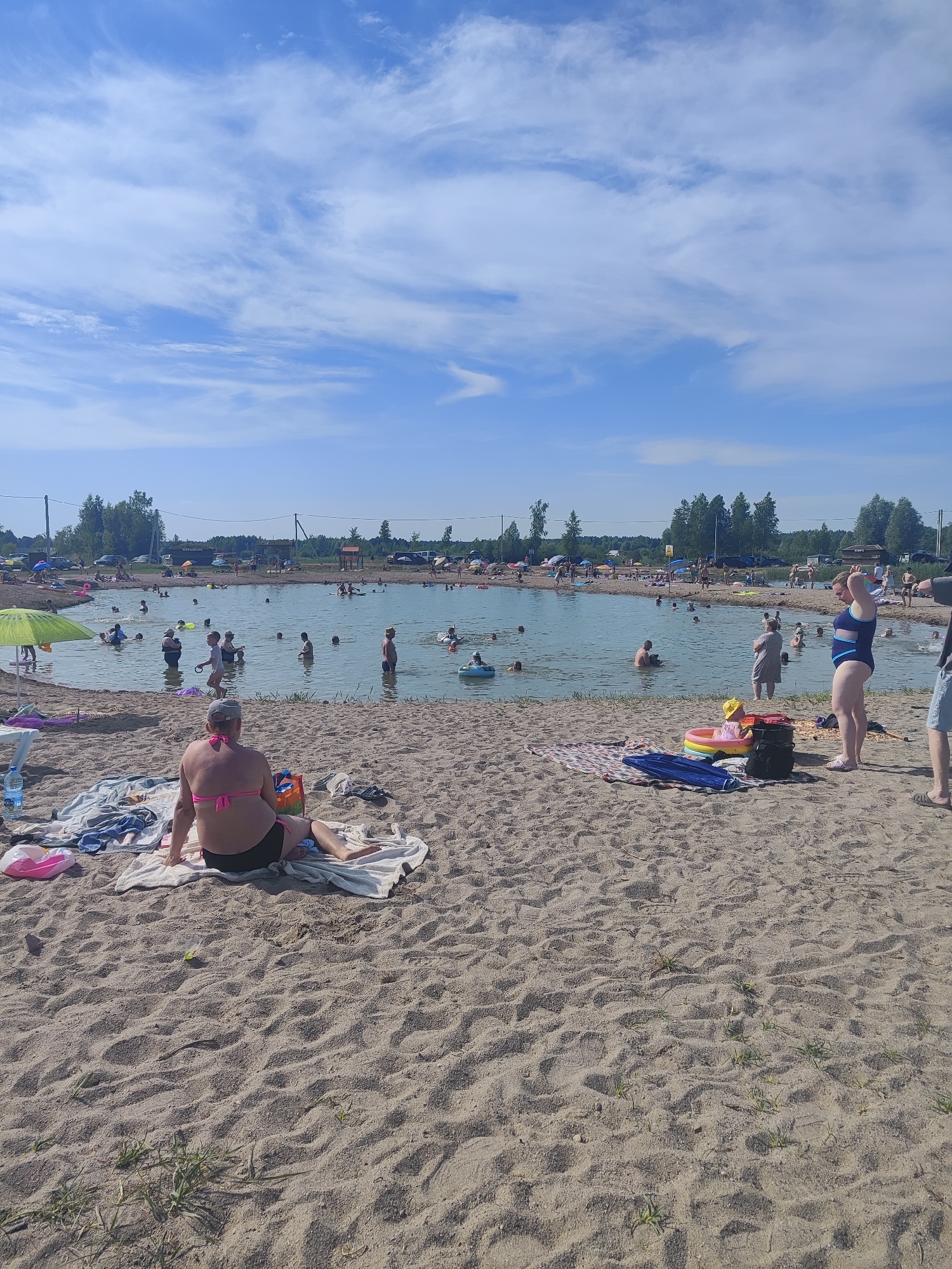 This image depicts a crowded beach scene on a sunny day, likely in Rovnoe, Russia. The primary subjects are numerous people enjoying a swim or relaxing on the sand. In the foreground, a woman in a pink bikini sits on a towel, facing away from the camera. To her right, a woman in a blue and white swimsuit stands looking out at the water.
The scene is centered around a body of water, possibly a lake or a large pond, where many individuals are swimming, wading, or playing. Some are using inflatable toys, including a child in a yellow hat in a small inflatable ring. The beach itself is sandy, with several towels and beach accessories scattered about. In the background, trees and some buildings or structures are visible, suggesting a recreational area. The sky is a clear blue with scattered white clouds, indicating fair weather and a bright, sunny day, likely mid-day.
The overall atmosphere appears to be one of leisure and enjoyment, with families and groups of people engaging in typical summer beach activities. The presence of numerous parked cars on the left side of the image suggests this is a popular local spot. No discernible text is visible in the image.