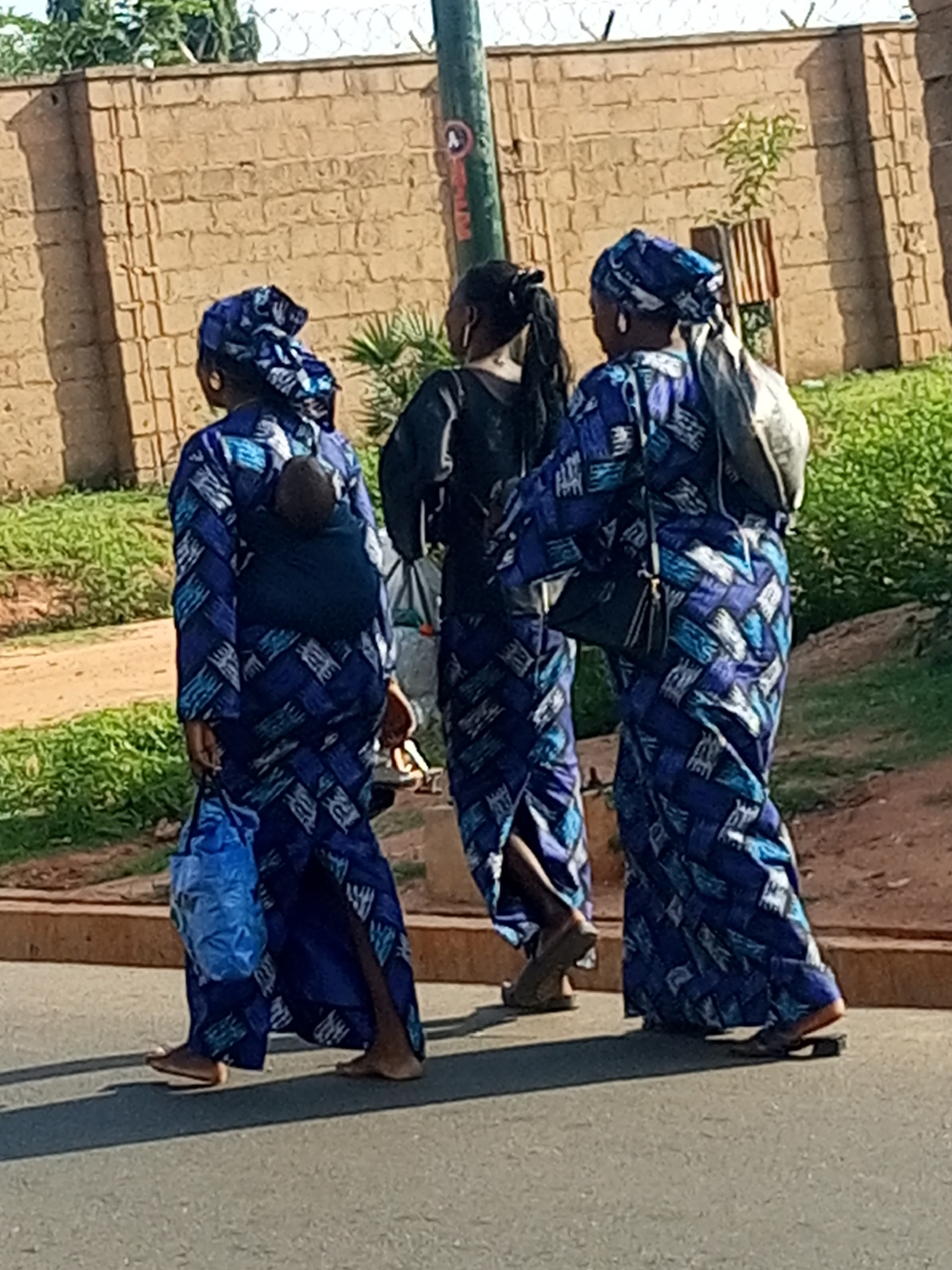 Three individuals are walking on a paved road, with a textured brick wall and greenery visible in the background. The individual on the left, facing away from the camera, wears a blue and white patterned garment and a matching headscarf. A baby is wrapped and carried on their back. This person is holding a blue plastic bag and wearing brown flat shoes. The person in the middle, also facing away, wears a black top and dark trousers. The individual on the right, facing away, wears a blue and white patterned garment and a matching headscarf. This person is carrying a dark bag over their shoulder and is wearing dark flat shoes. The scene appears to be outdoors on a sunny day. The location is identified as Tempure, Nigeria.