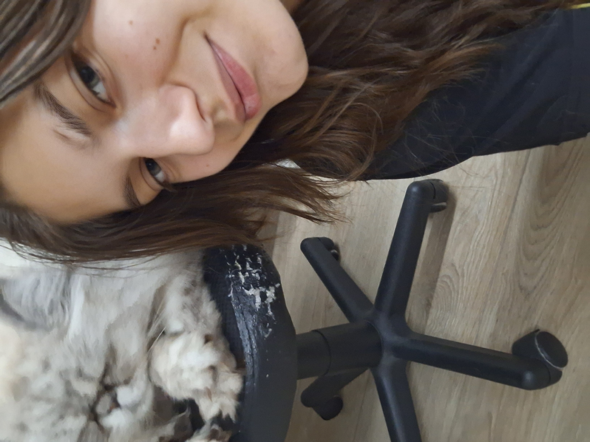 A selfie taken indoors, rotated counter-clockwise. A young woman with light skin, brown hair, and a slight smile is prominently featured. To her left is a fluffy, light-colored animal, possibly a cat or dog, resting on a chair. A black office chair with a five-star base sits on a light-colored wooden floor. The scene appears to be in a domestic setting. There is no information available about the location being in Panagyurishte, Bulgaria.